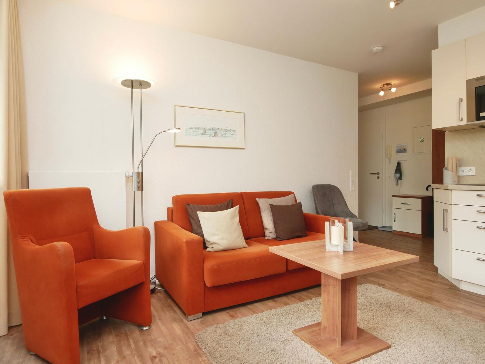 Wohnzimmer mit orangefarbenem Sofa und Sessel, Holztisch und Küchenbereich im Hintergrund.
