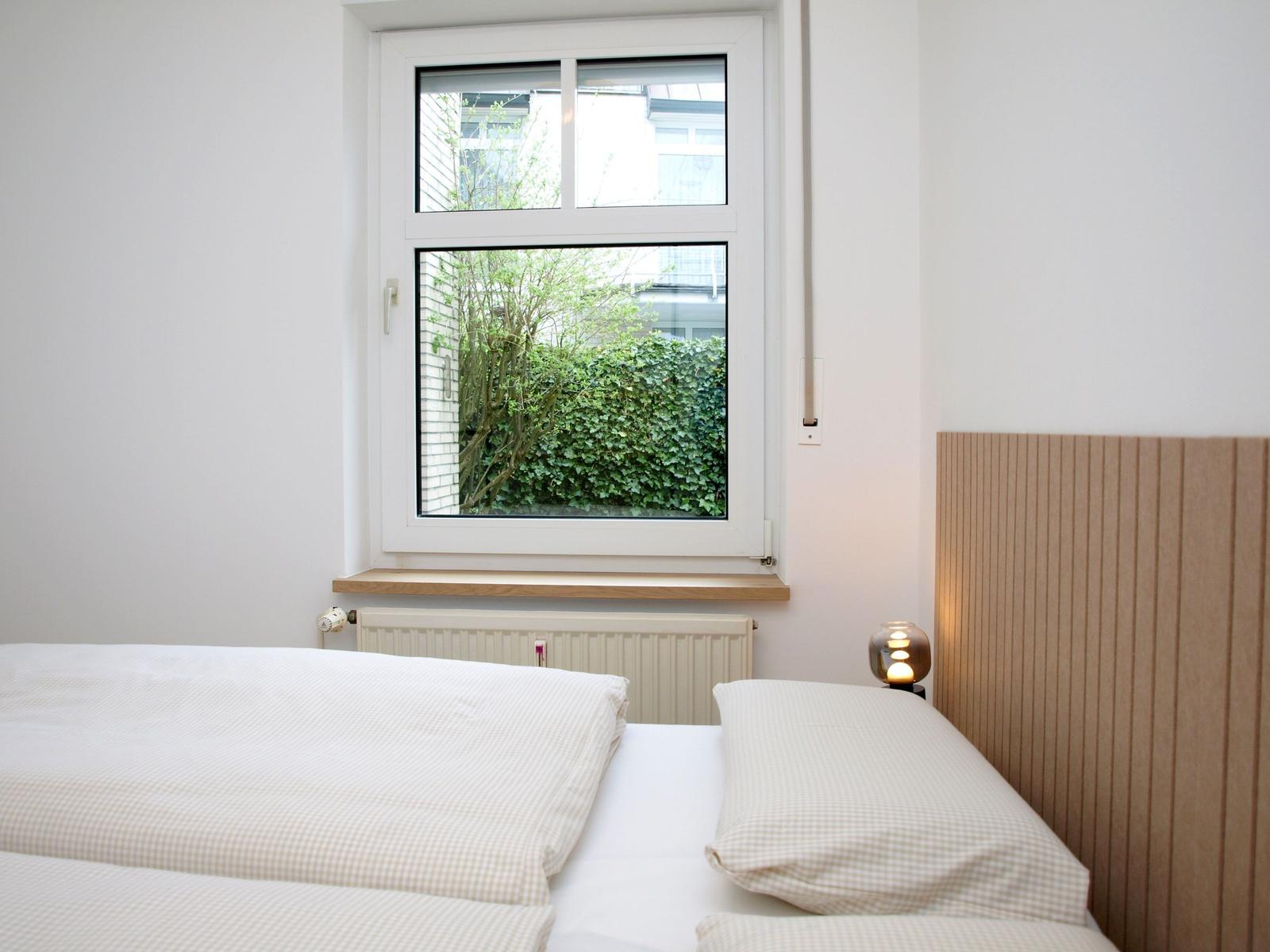 Ein Bett mit Holzkopfteil und Fensterblick auf grüne Pflanzen.