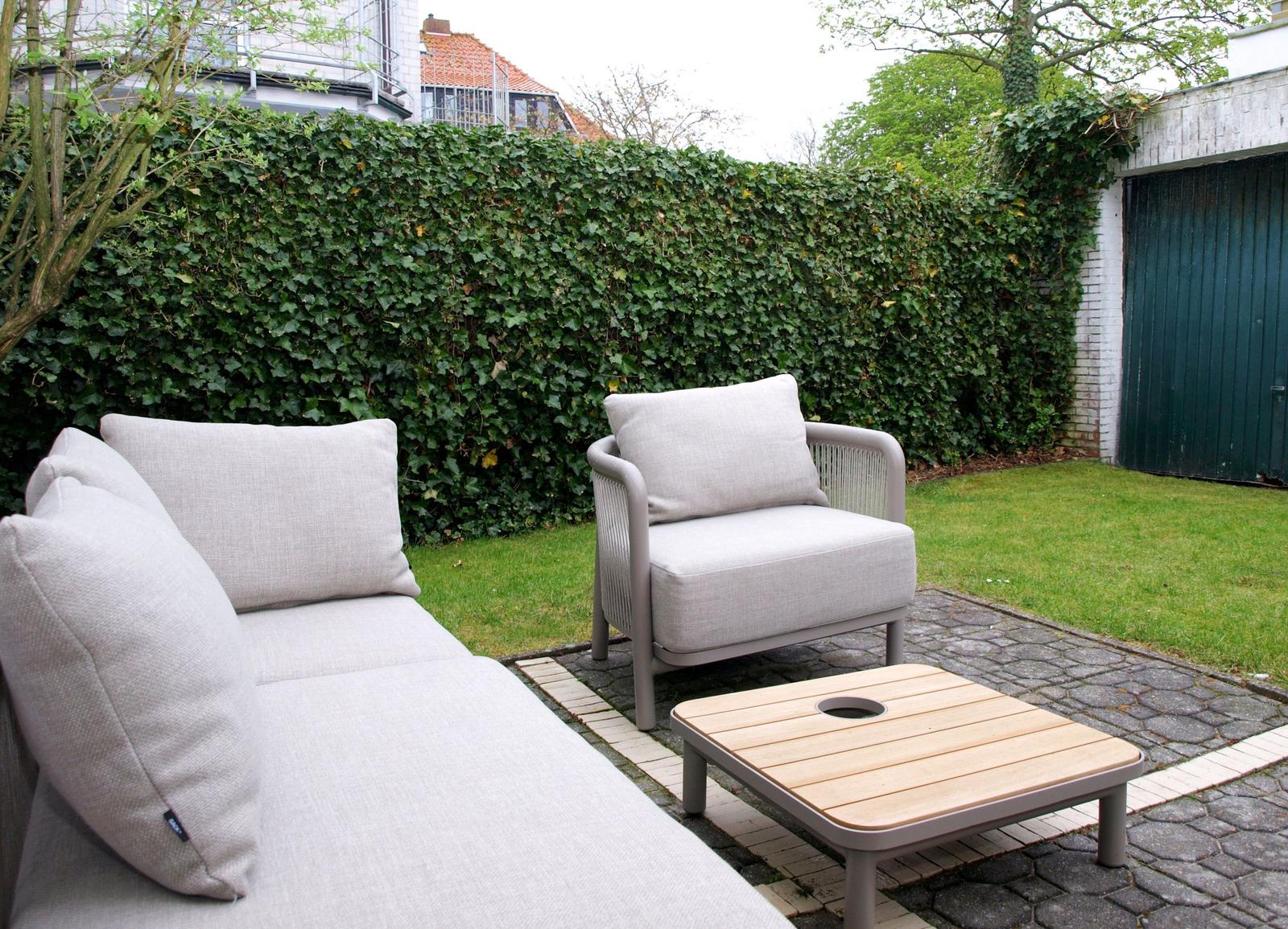 Außenbereich mit Sofa, Sessel und Tisch auf gepflastertem Boden vor grüner Hecke.