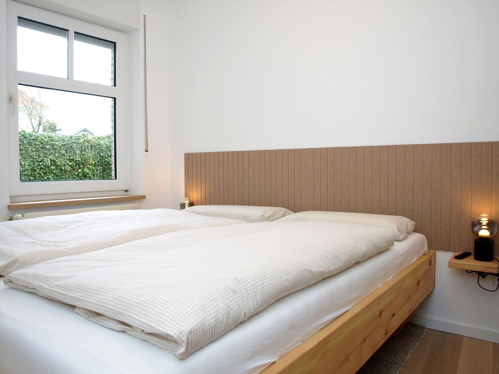 Schlafzimmer mit Doppelbett, Holzgestell und Holzpaneelen an der Wand. Fenster mit Blick auf grüne Hecke.