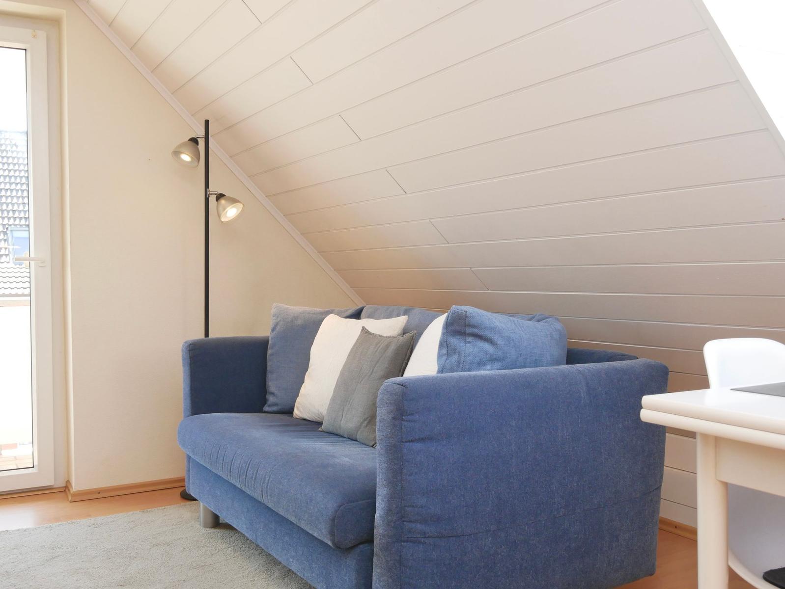 Blauer Sofa mit Kissen unter schrägem Dach mit Stehleuchte und Fenster.
