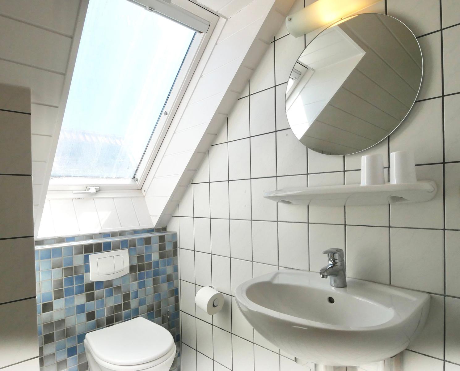 Badezimmer mit Waschbecken, Spiegel und Dachfenster. Wandfliesen und Toilette sind sichtbar.
