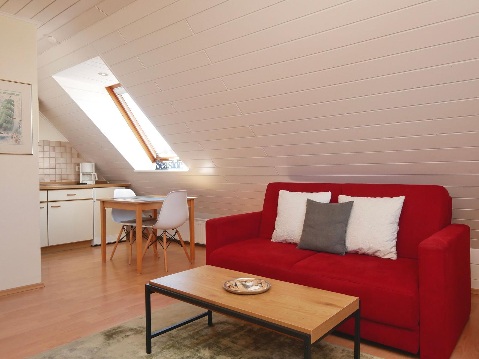 Ein Wohnbereich mit rotem Sofa, Holztisch und Dachschrägen. Küchenbereich mit Tisch und Stühlen.