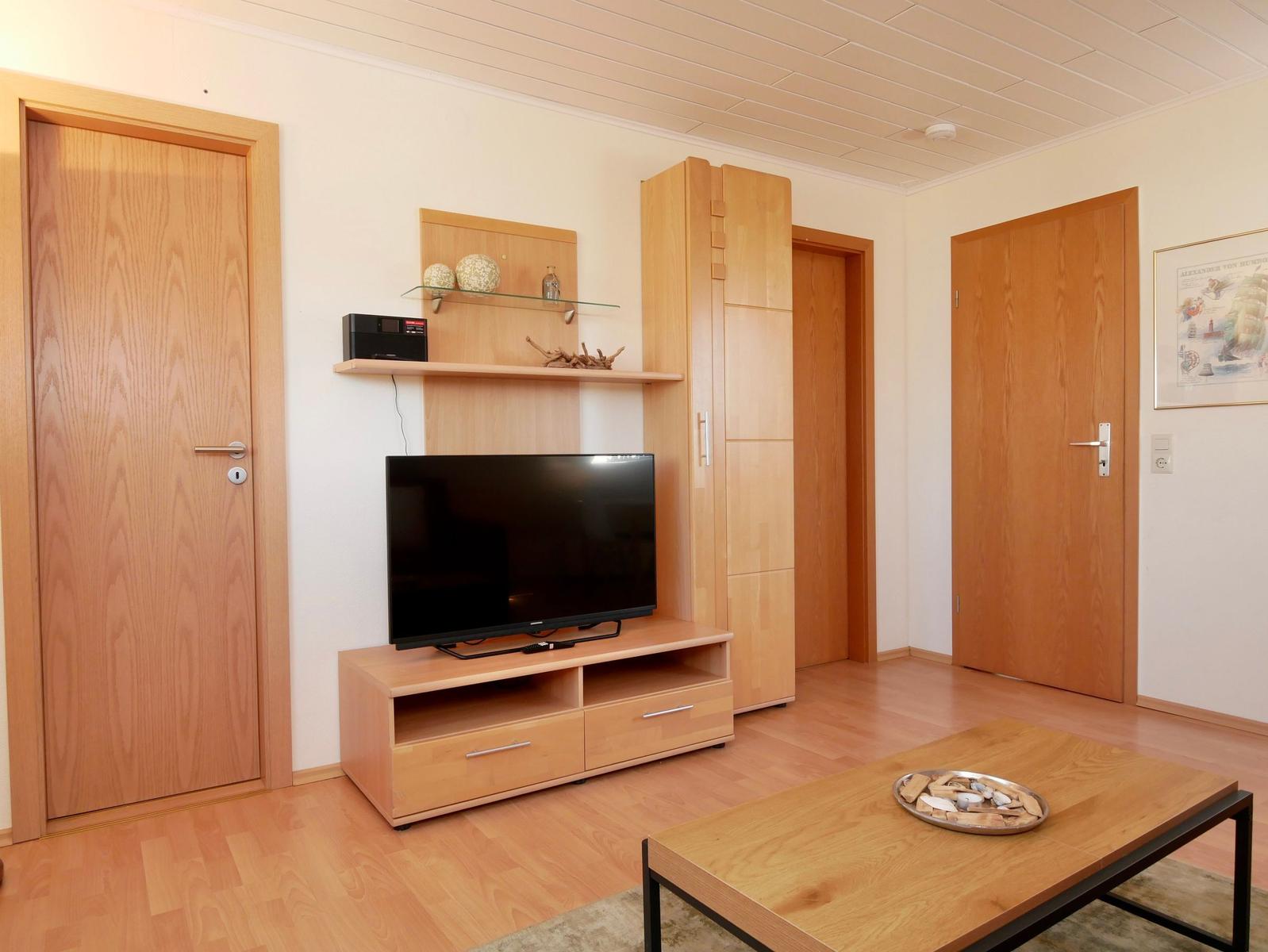 Wohnzimmer mit Fernseher, Holzmöbeln und Tisch. Mehrere Türen und Wanddekoration sichtbar.
