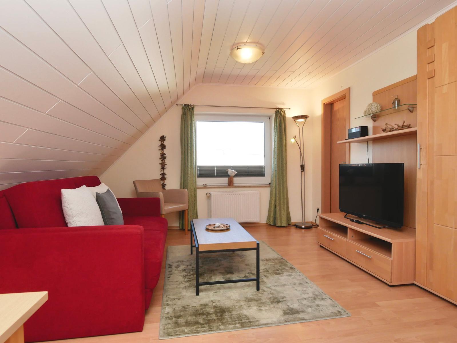 Ein Wohnzimmer mit rotem Sofa, TV und Holzmöbeln unter einer schrägen Decke.