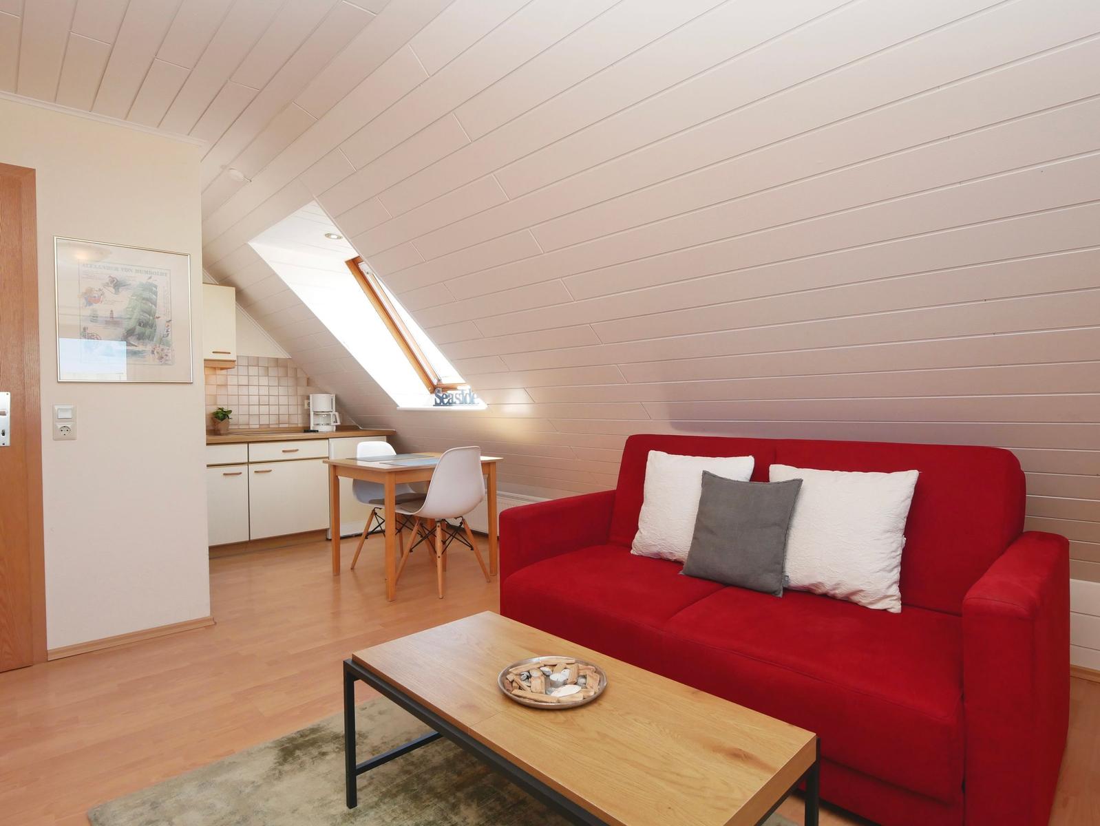 Ein Wohnbereich mit rotem Sofa, Holztisch und schrägem Dach. Im Hintergrund Küche und Tisch.