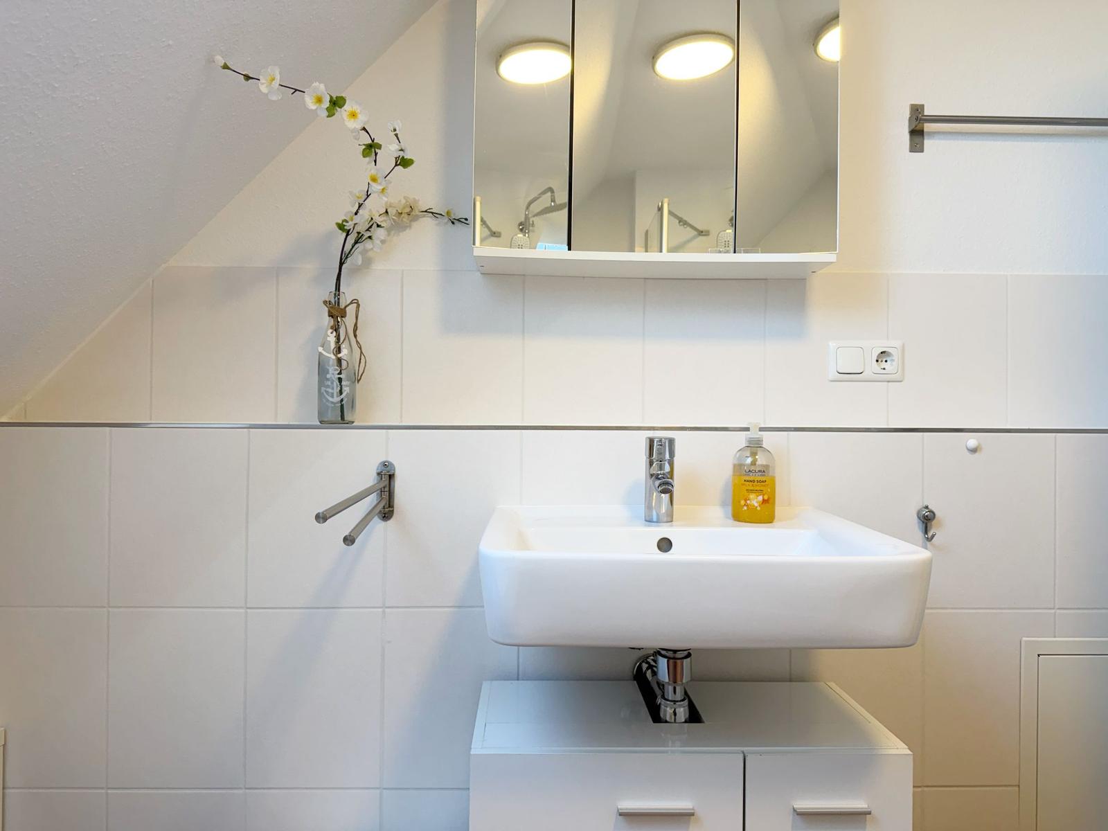 Waschbecken mit Spiegel, Handseife und Blumenvase an der Wand.