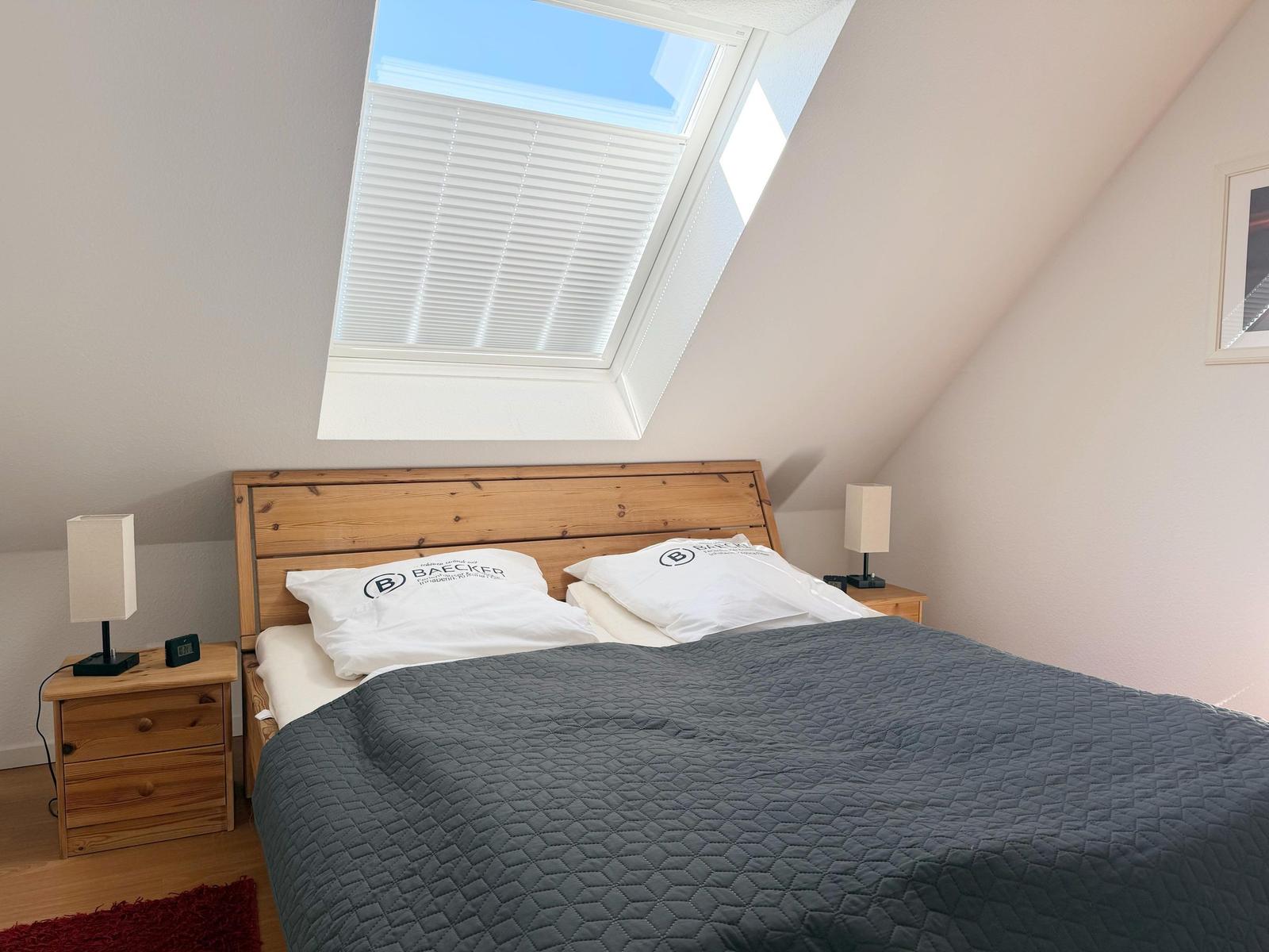 Schlafzimmer mit Dachschrägen, Bett, Nachttisch und Dachfenster mit Jalousie.