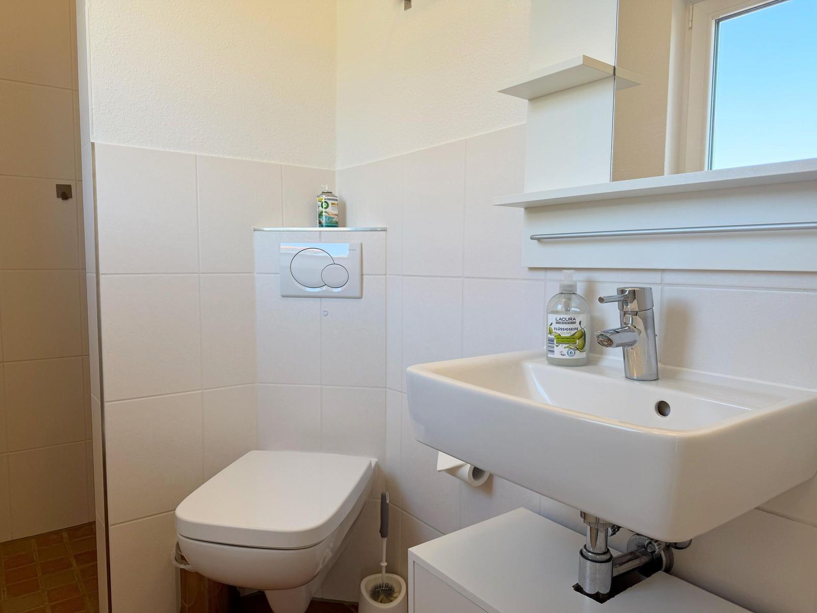 Badezimmer mit Toilette, Waschbecken und Spiegel. Fliesenwand und Fenster.