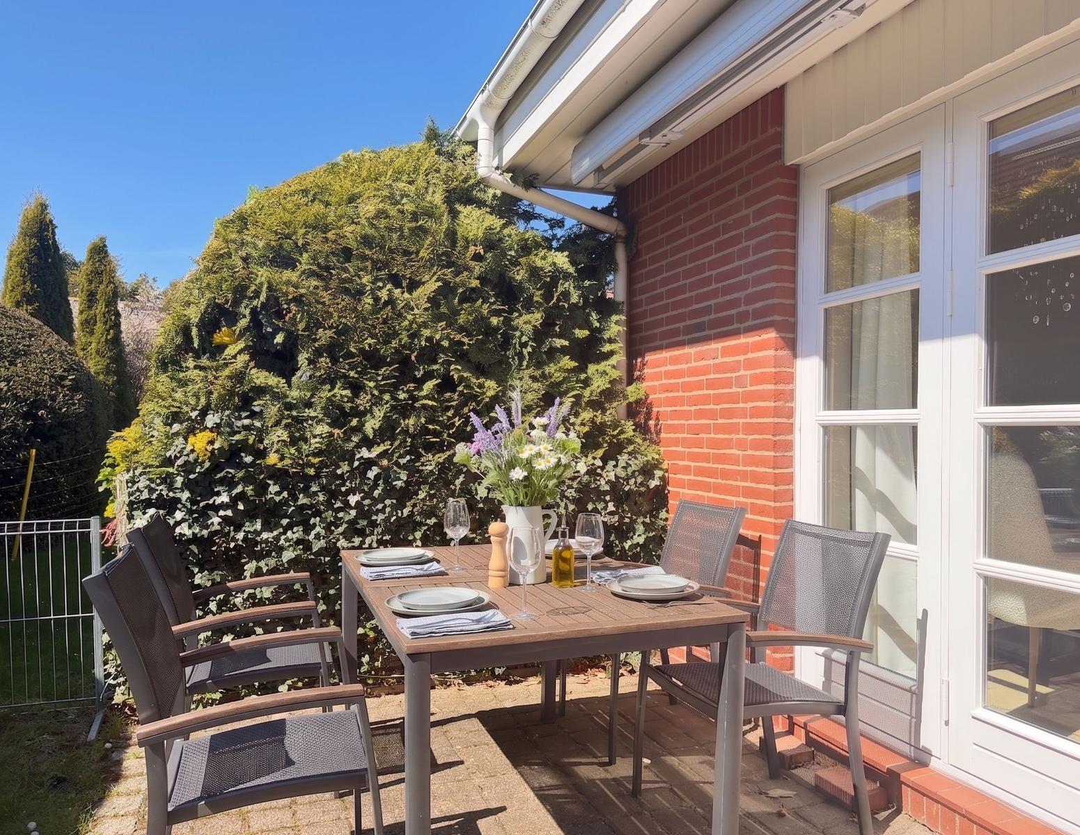 Terrasse mit Tisch, Stühlen und Blumenarrangement vor rotem Backsteinhaus.