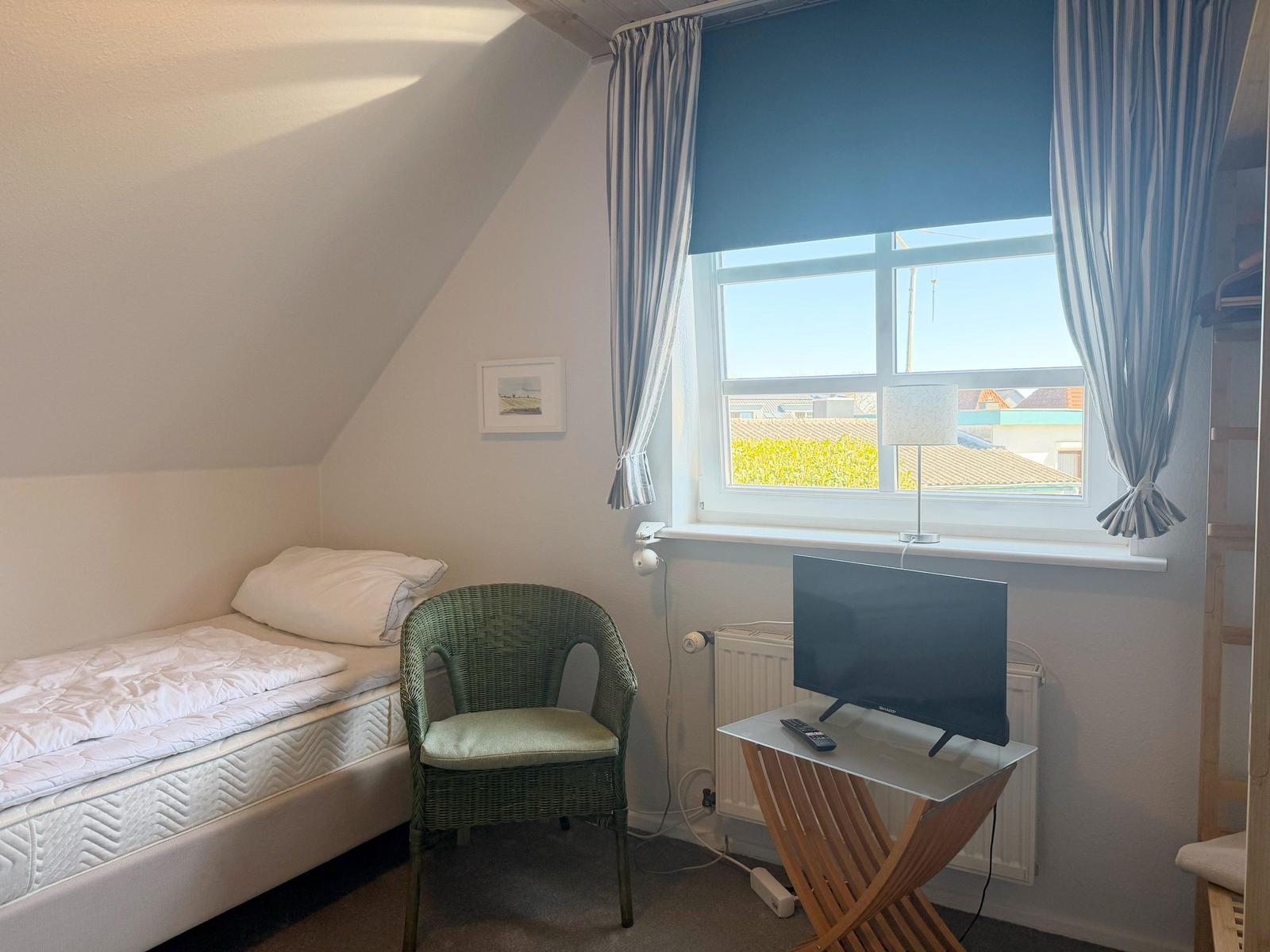 Ein Schlafzimmer mit Dachschrägen, Bett, Stuhl und TV. Fenster mit Vorhängen.