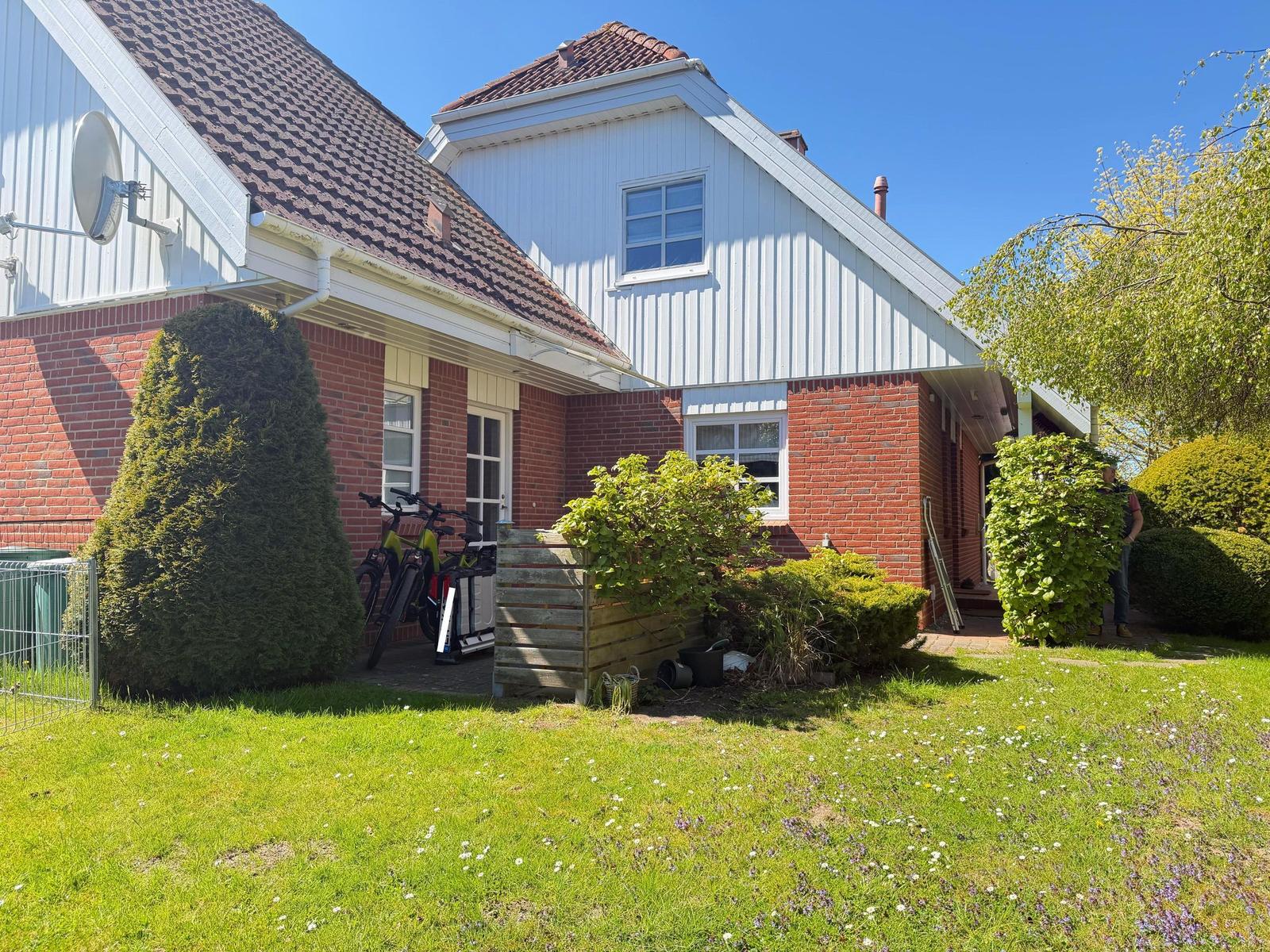 Haus mit rotem Ziegelunterbau und weißem Holzobergeschoss, begrünter Vorgarten mit Fahrrädern.