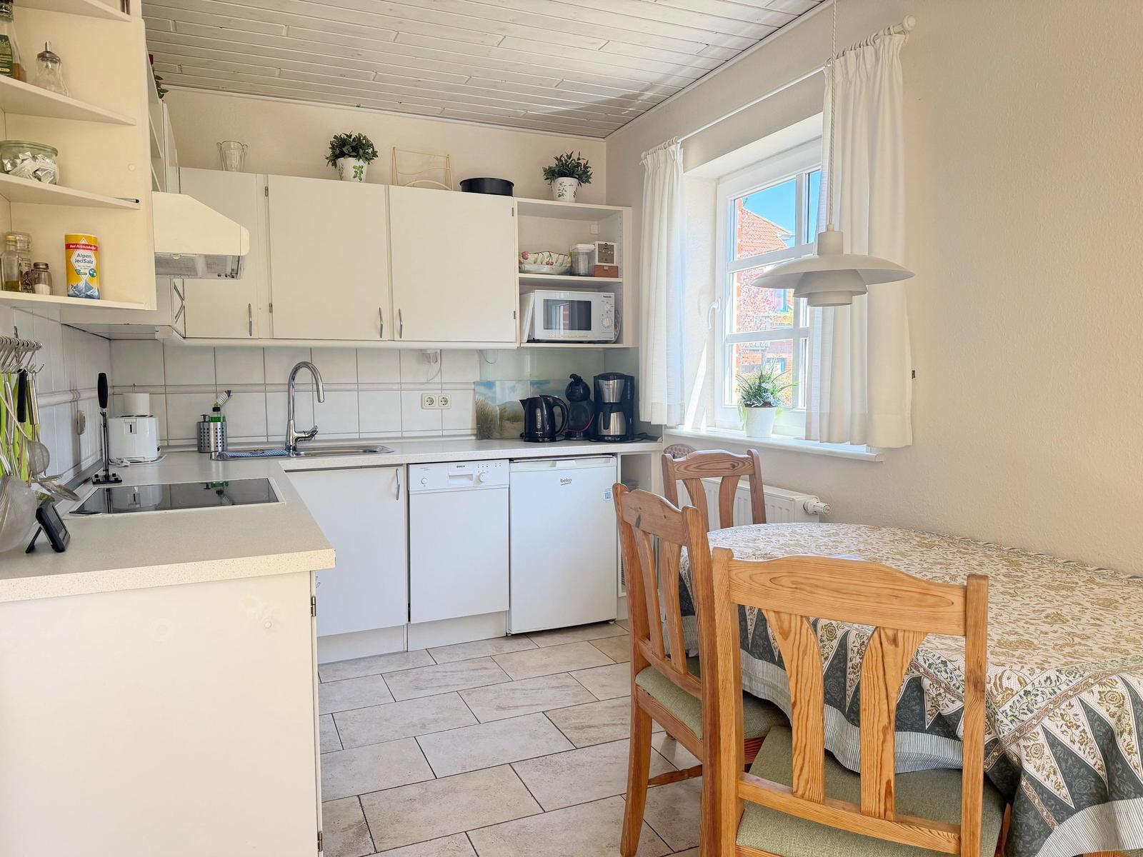 Küche mit Essbereich, weißen Schränken und Holzstühlen. Fenster mit Vorhängen und Tischdecke.