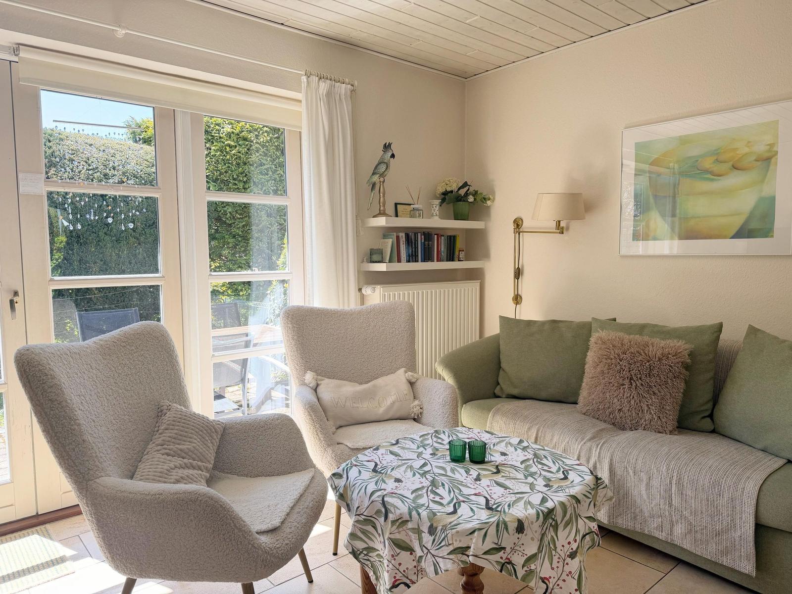 Ein Wohnzimmer mit Sofa, zwei Sesseln und Tisch. Durch Fenster ist Garten sichtbar.