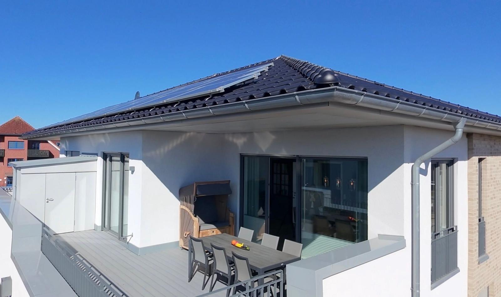 Terrasse mit Tisch, Stühlen und Liegestuhl. Dach mit Solarpaneelen. Modernes Haus mit Glasfront.