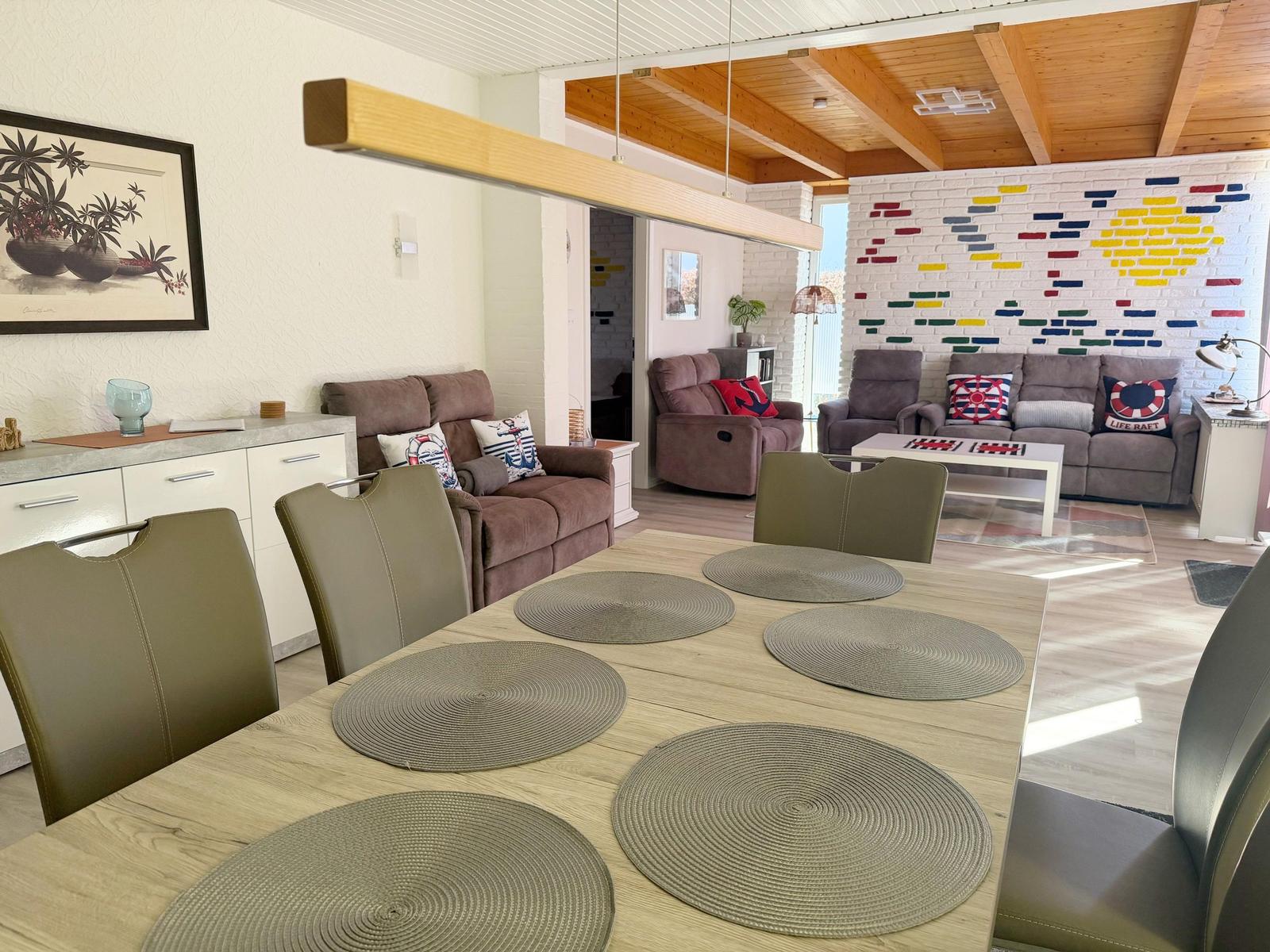 Essbereich mit Tisch und Stühlen. Im Hintergrund Wohnzimmer mit Sofa und dekorativer Wand.