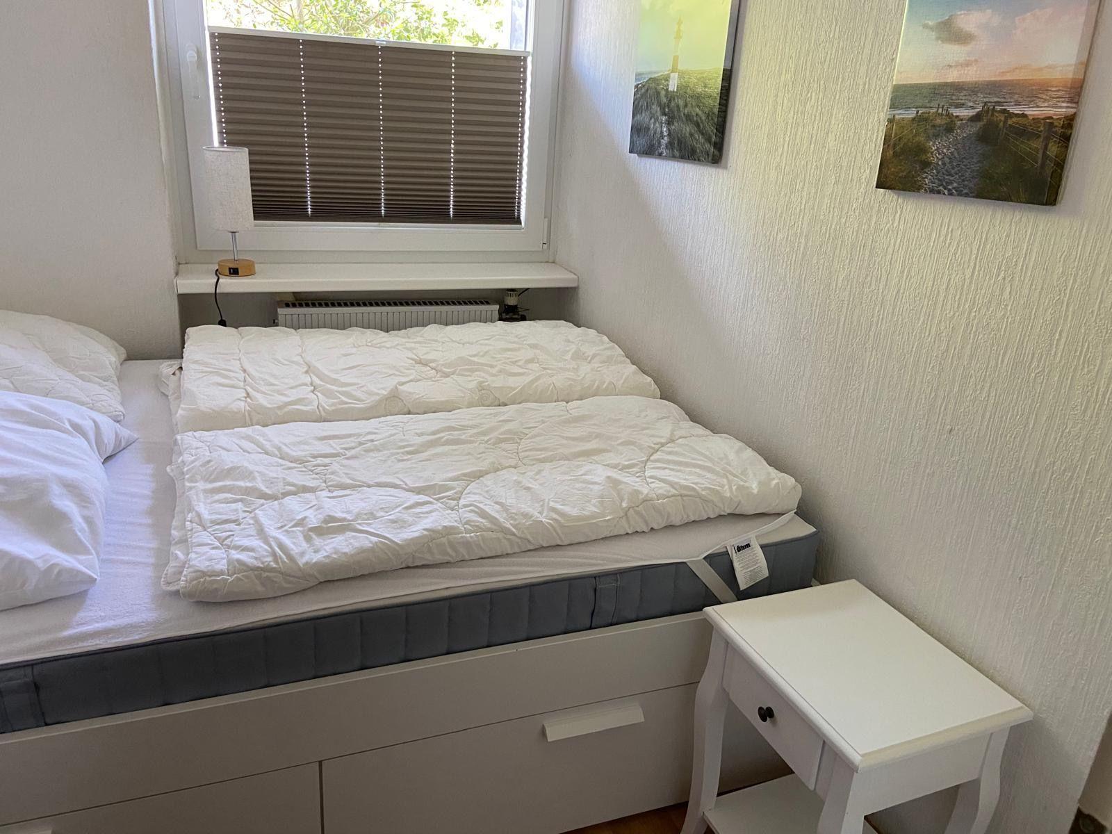 Schlafzimmer mit Bett, Nachttisch und Fenster mit Jalousie.