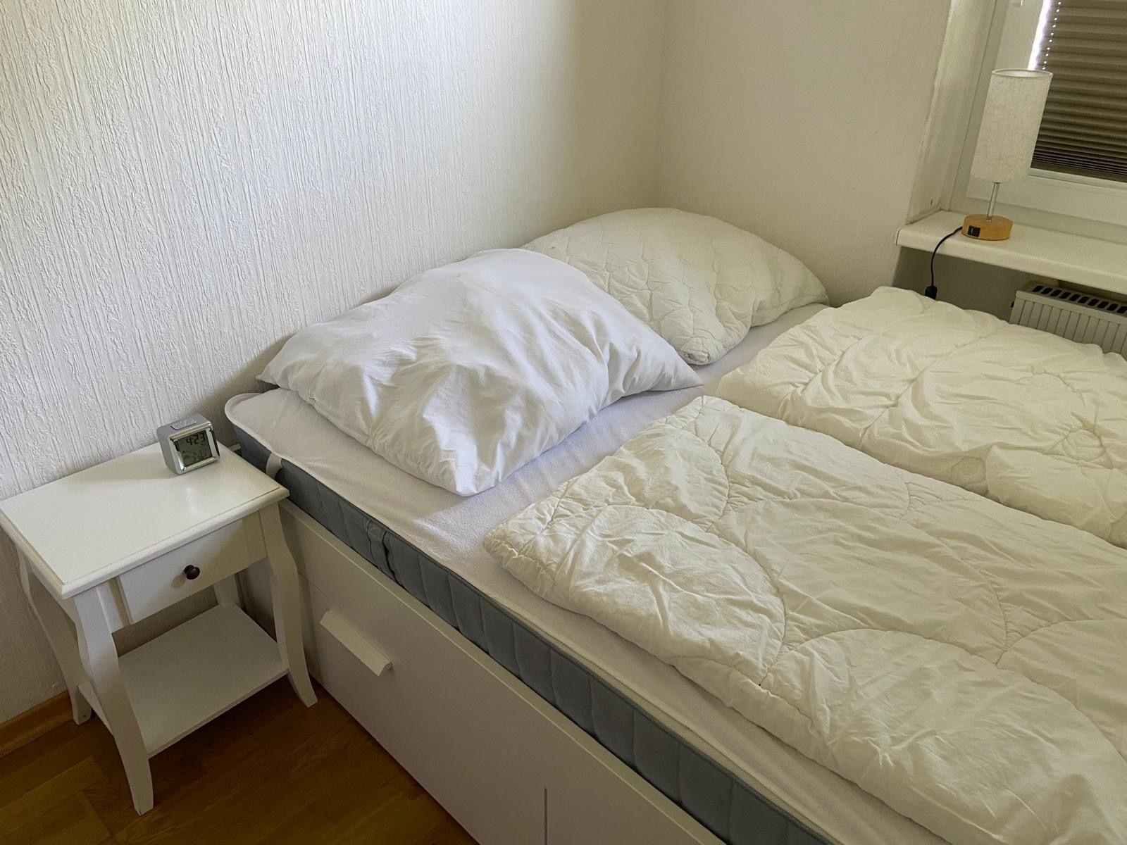 Schlafzimmer mit Bett, Nachttisch und Lampe an Fensterbank.