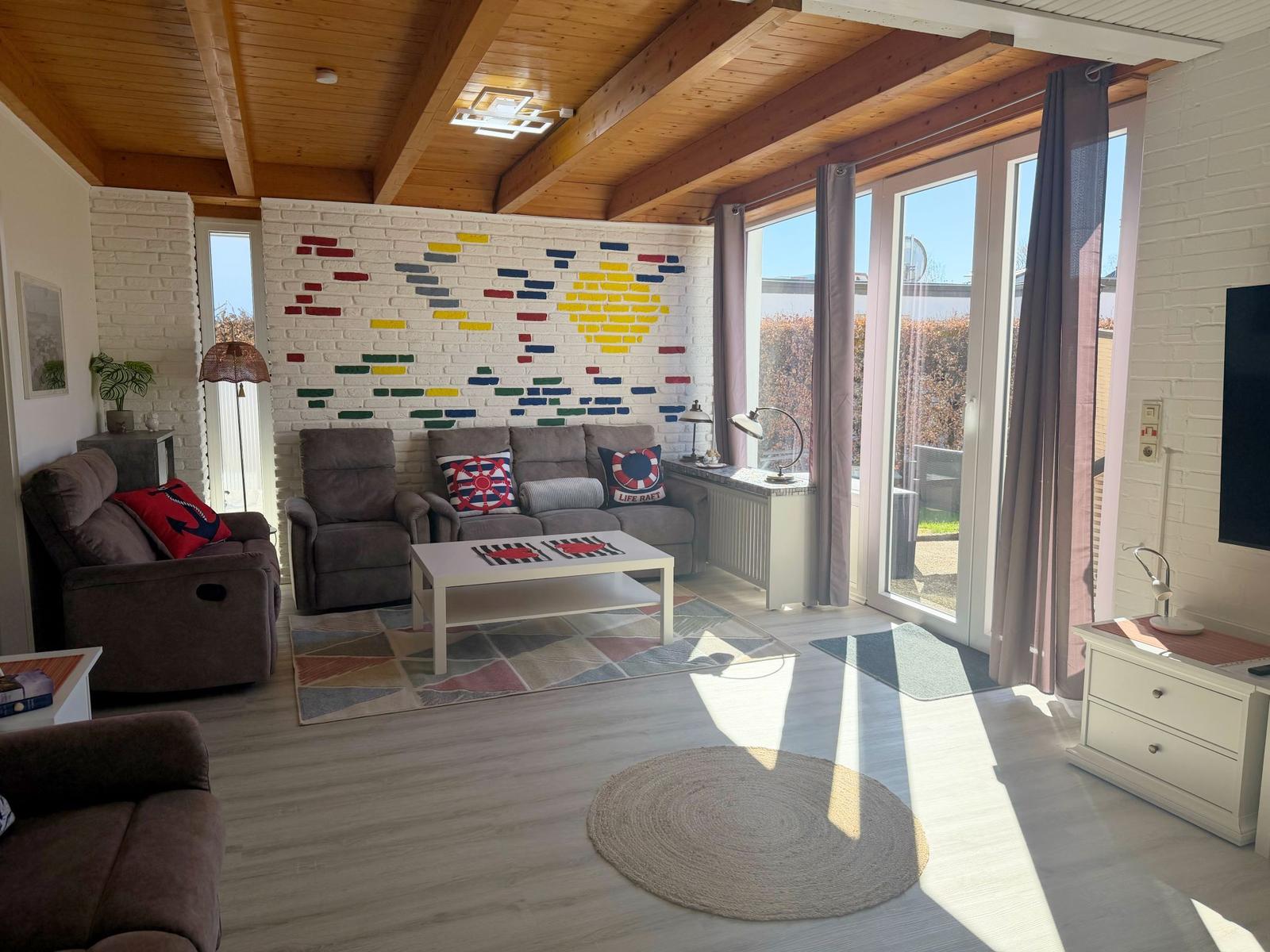 Wohnzimmer mit Sofa, Sessel und buntem Wandbild. Große Fenster und Holzdecke.