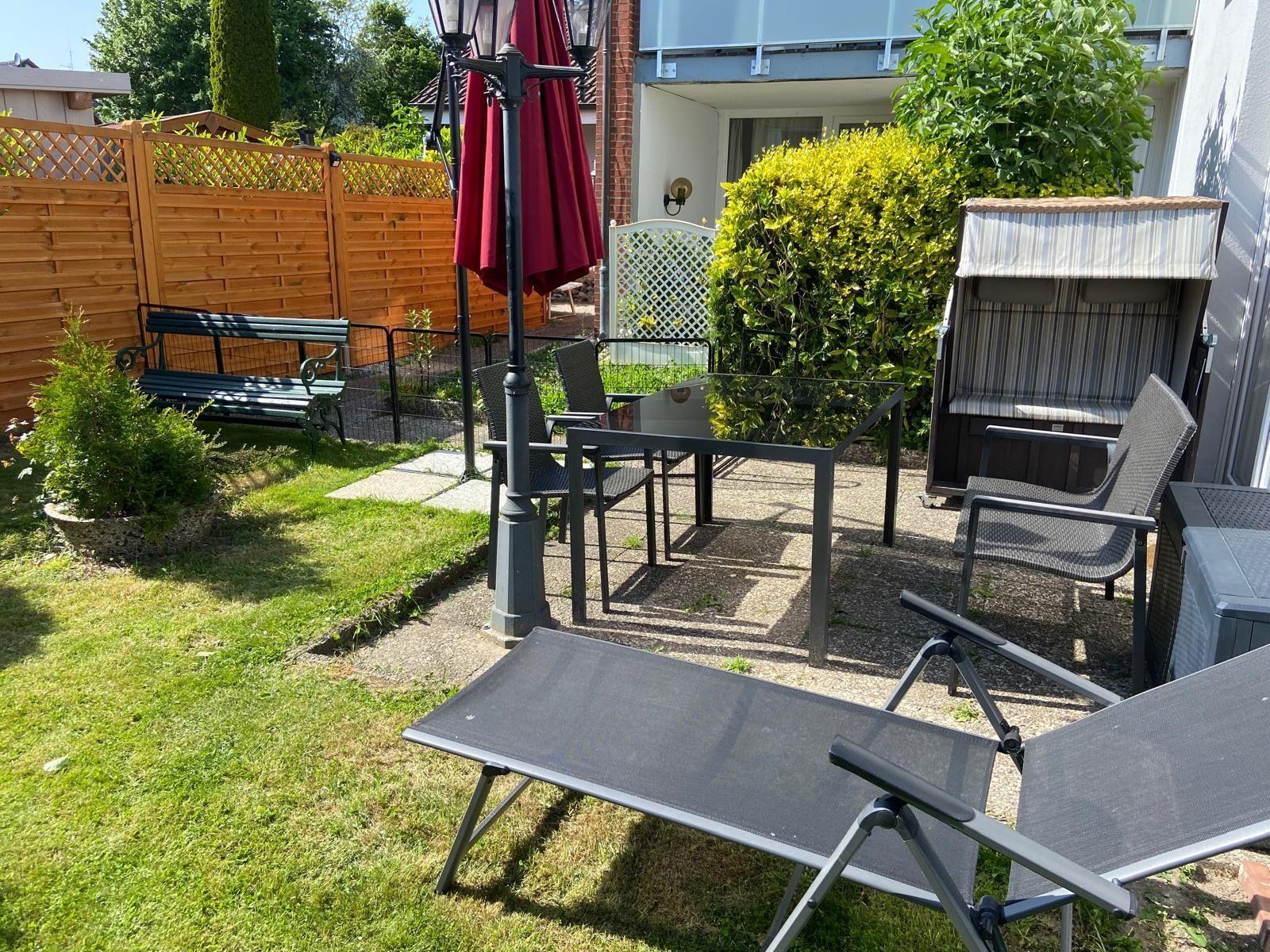 Hinterhof mit Gartenmöbeln, Liegestühlen und Sonnenschirm. Grasfläche und Holzzaun sichtbar.