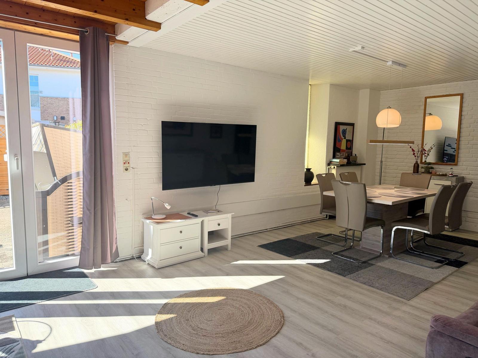 Wohn- und Essbereich mit TV, Tisch und Stühlen, Licht und Blick auf Balkon.