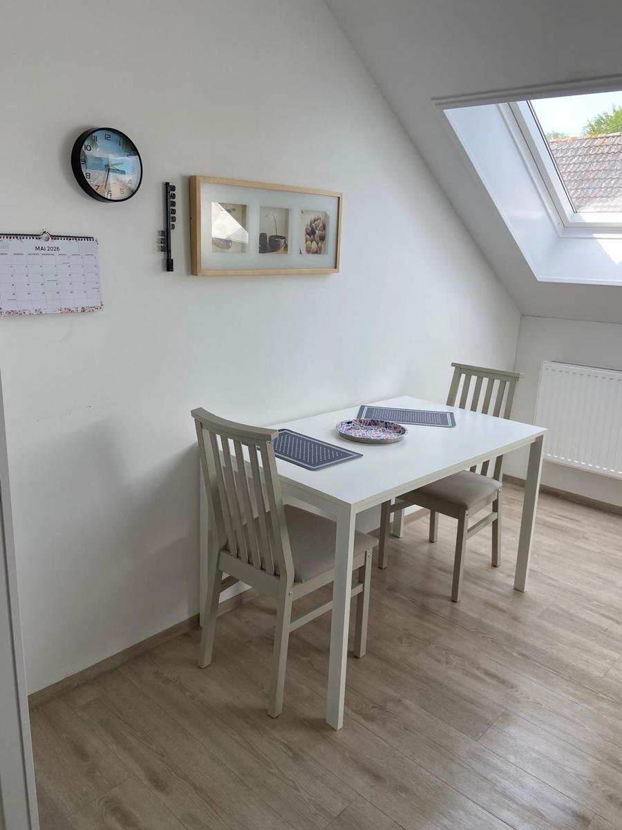Essbereich mit Tisch, zwei Stühlen, Wanduhr und Fenster. Holzdielenboden.