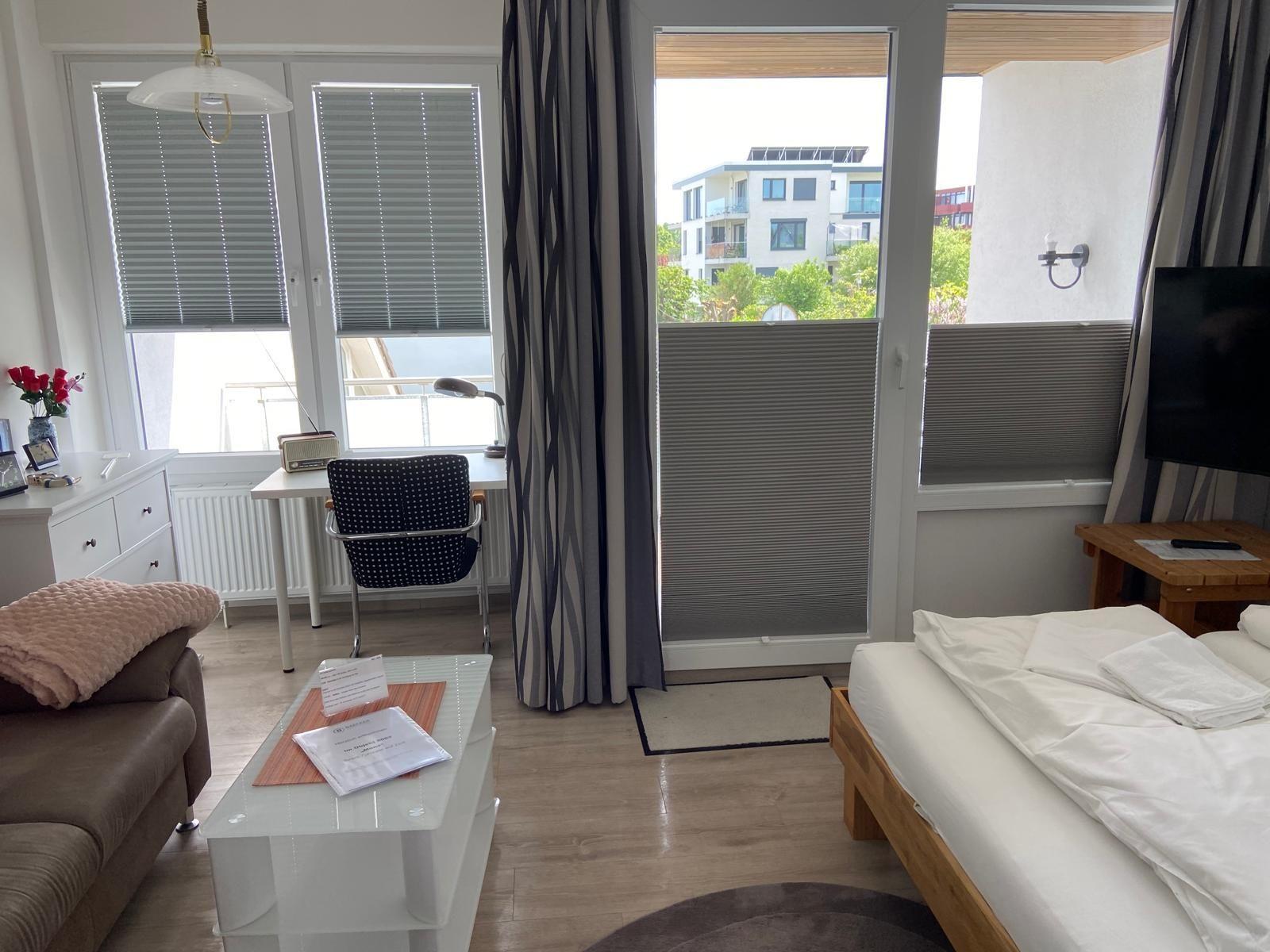 Zimmer mit Bett, Schreibtisch, Sofa und Balkon mit Blick auf Gebäude.