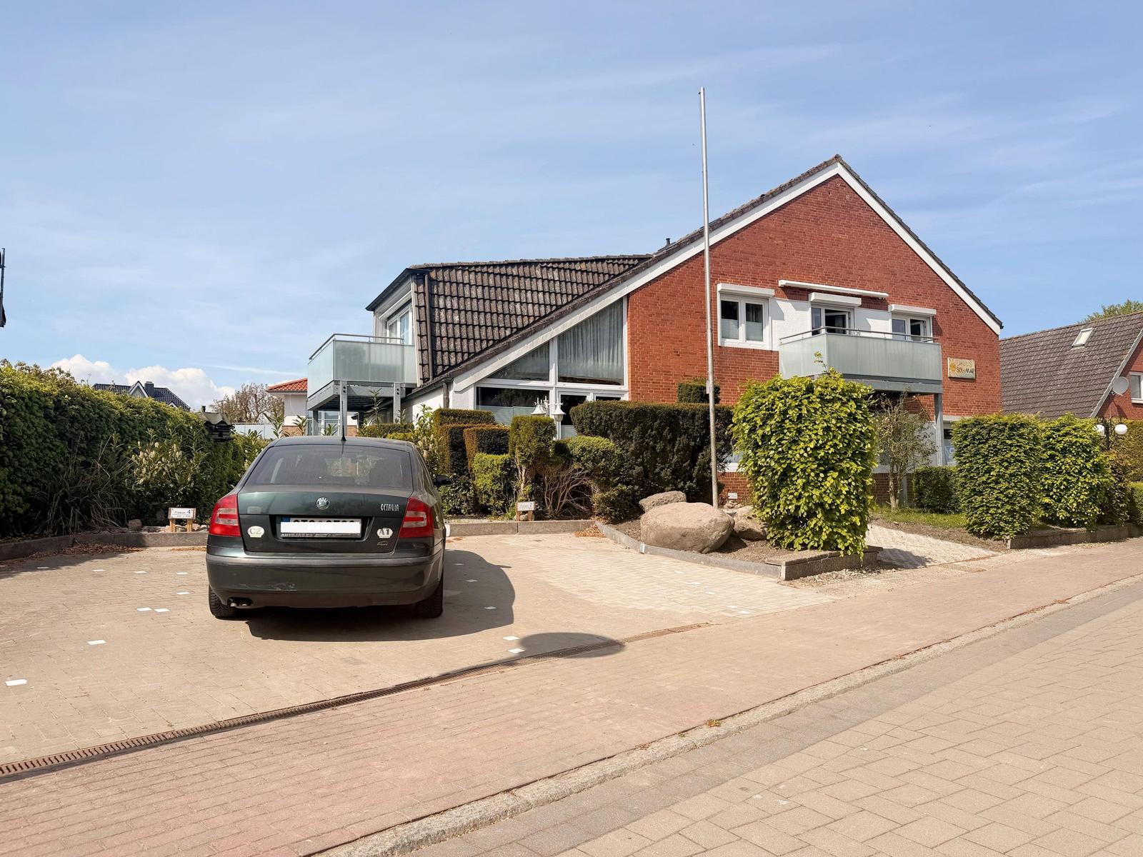 A two-story house with a balcony, surrounded by greenery and a parking area.