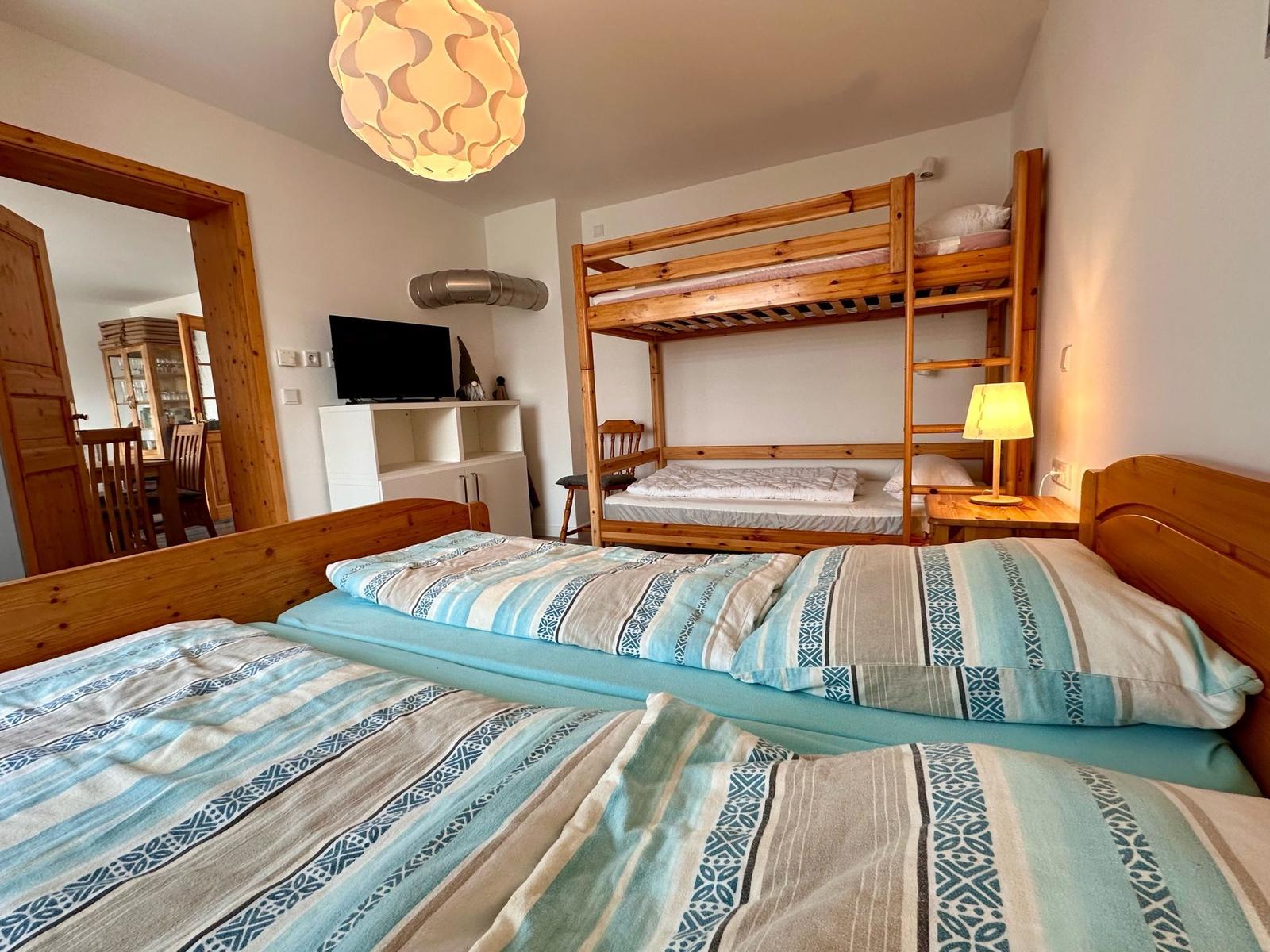 Zimmer mit zwei Einzelbetten und einem Holz-Kinderstockbett. Fernseher und Tischlampe sind vorhanden.