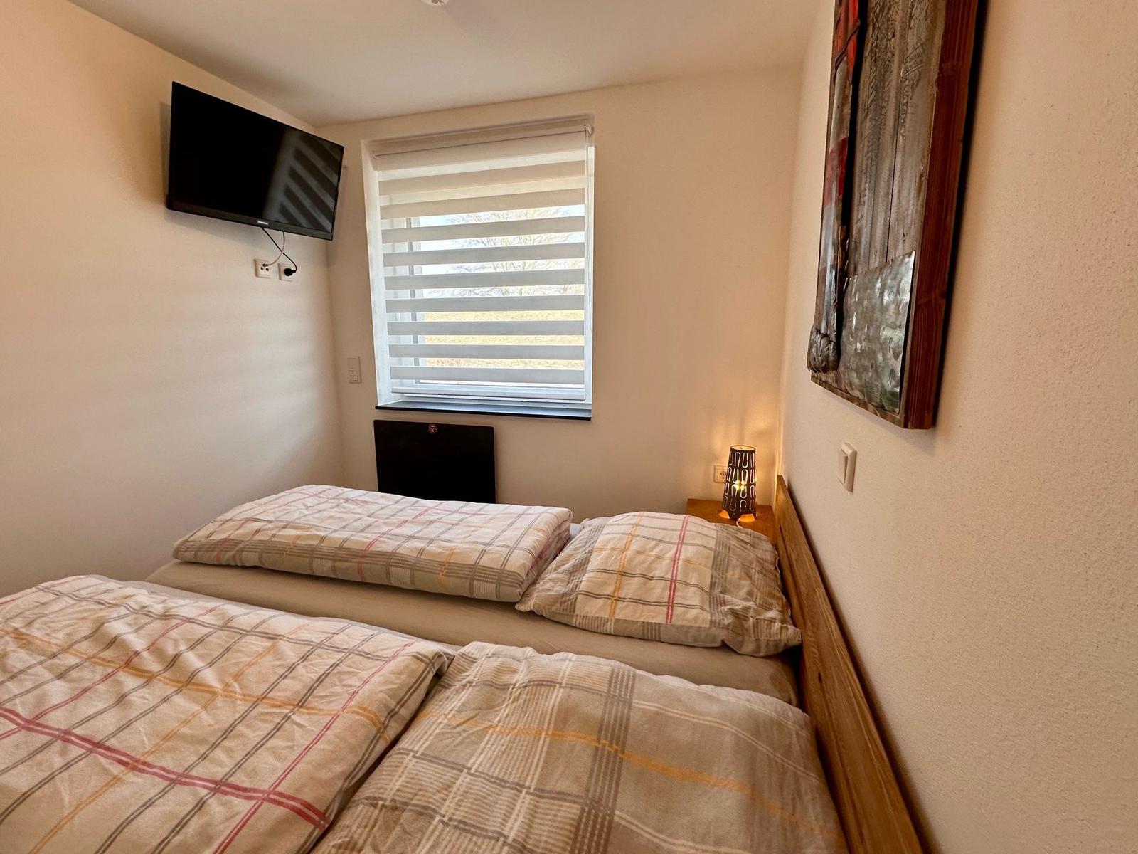 Schlafzimmer mit Doppelbett, Fenster, TV und Nachttisch mit Lampe.