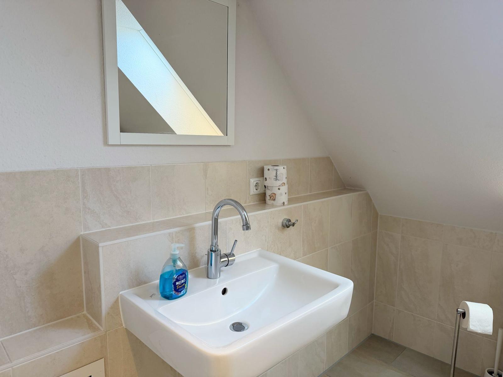 Badezimmer mit Waschbecken, Spiegel und Toilettenpapierhalter unter schräger Decke.