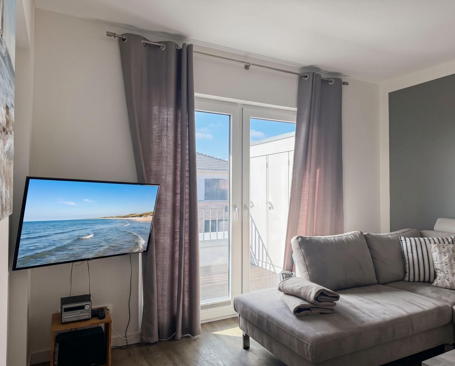 Wohnzimmer mit Sofa, TV und Balkontür. Vorhang und Holzboden sichtbar.