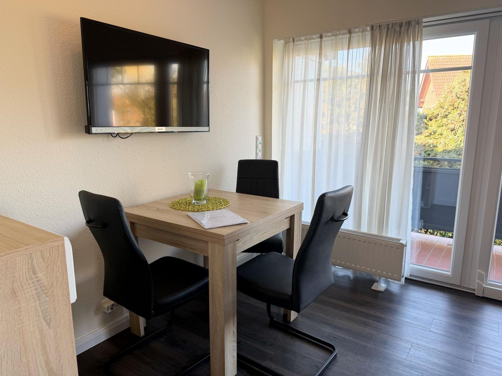 Essbereich mit Tisch, drei Stühlen, Fernseher und Balkontür.