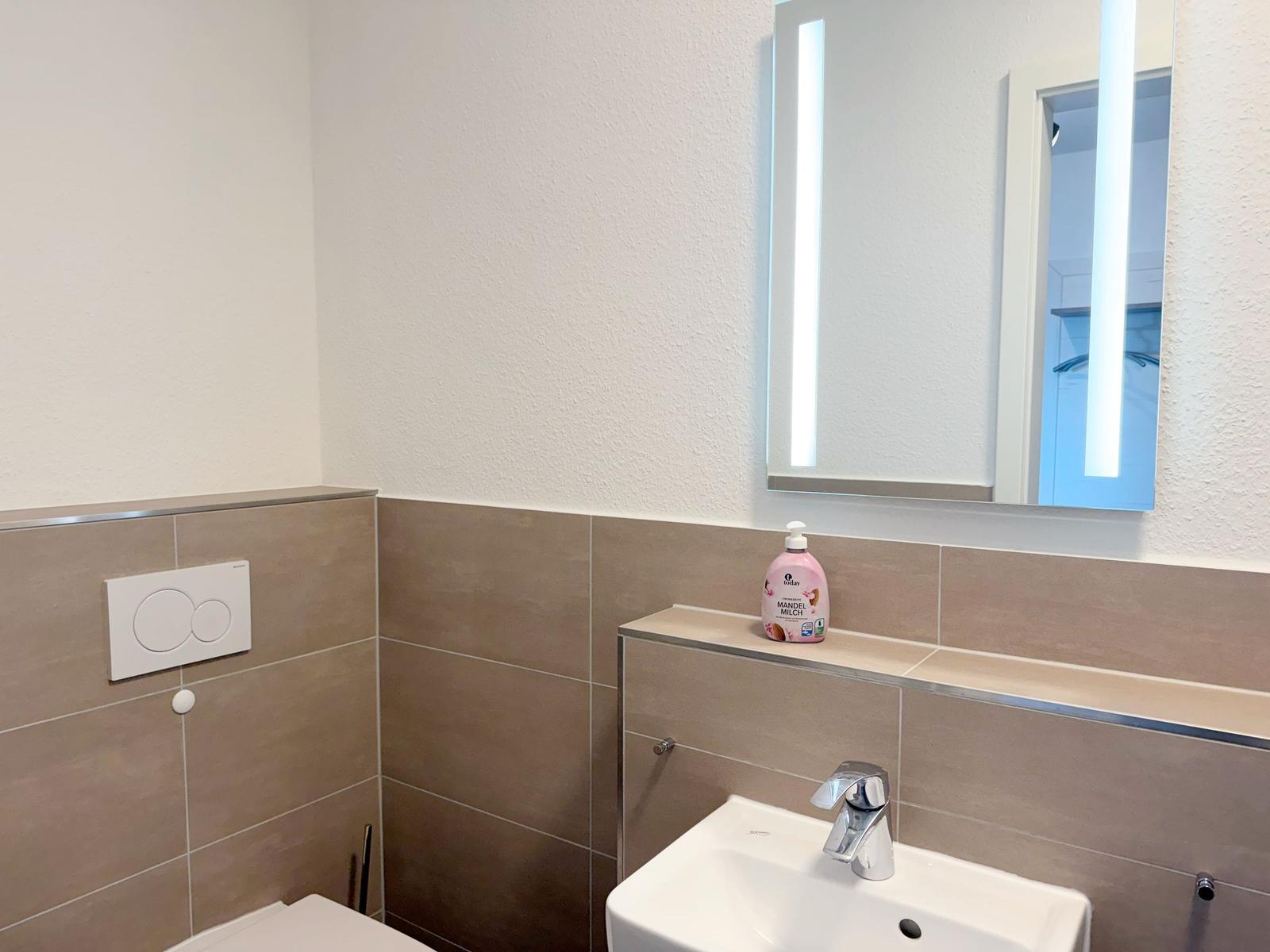 Badezimmer mit Waschbecken, Spiegel und Toilette. Auf der Ablage steht eine Flasche.