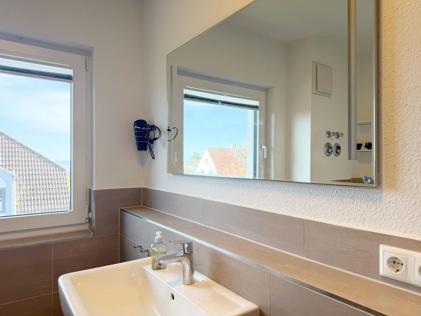 Badezimmer mit Waschbecken, Spiegel und Fenster mit Blick auf Dächer.