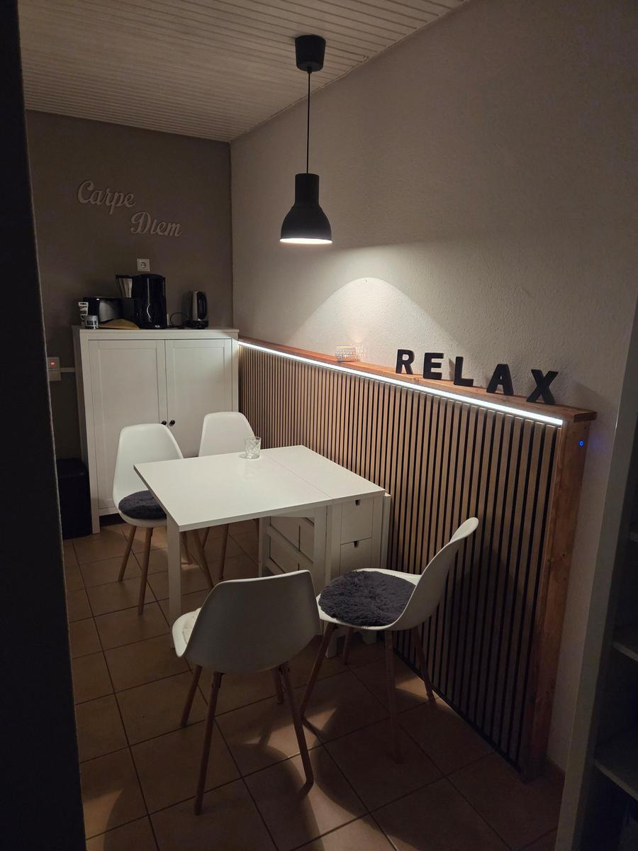 Essbereich mit Tisch, Stühlen und Wanddekoration. Hintergrund: Küchenzeile mit Kaffeemaschine.