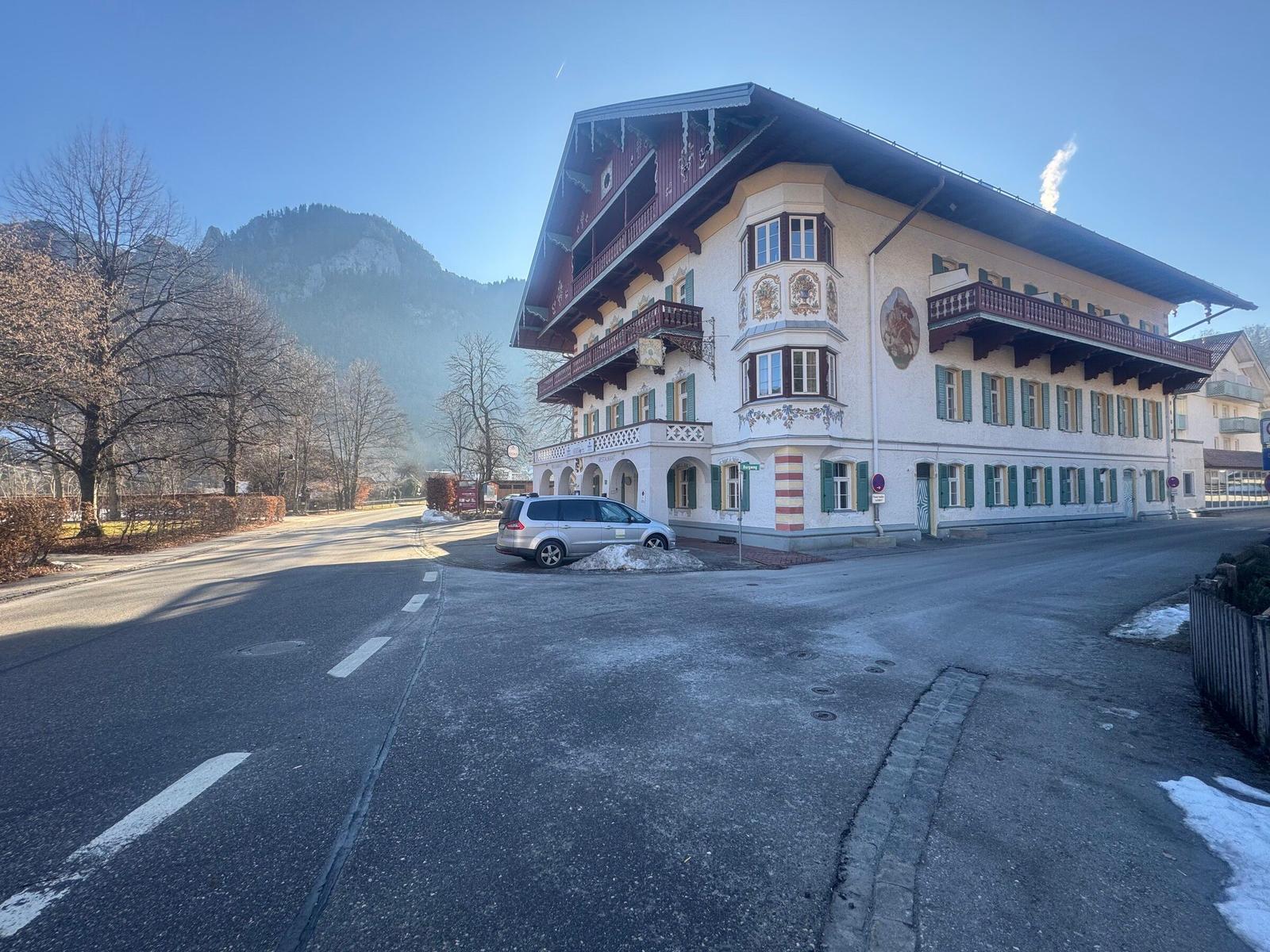 Altes Gebäude mit Balkonen und grünen Fensterläden an einer Straße mit Bergen im Hintergrund.