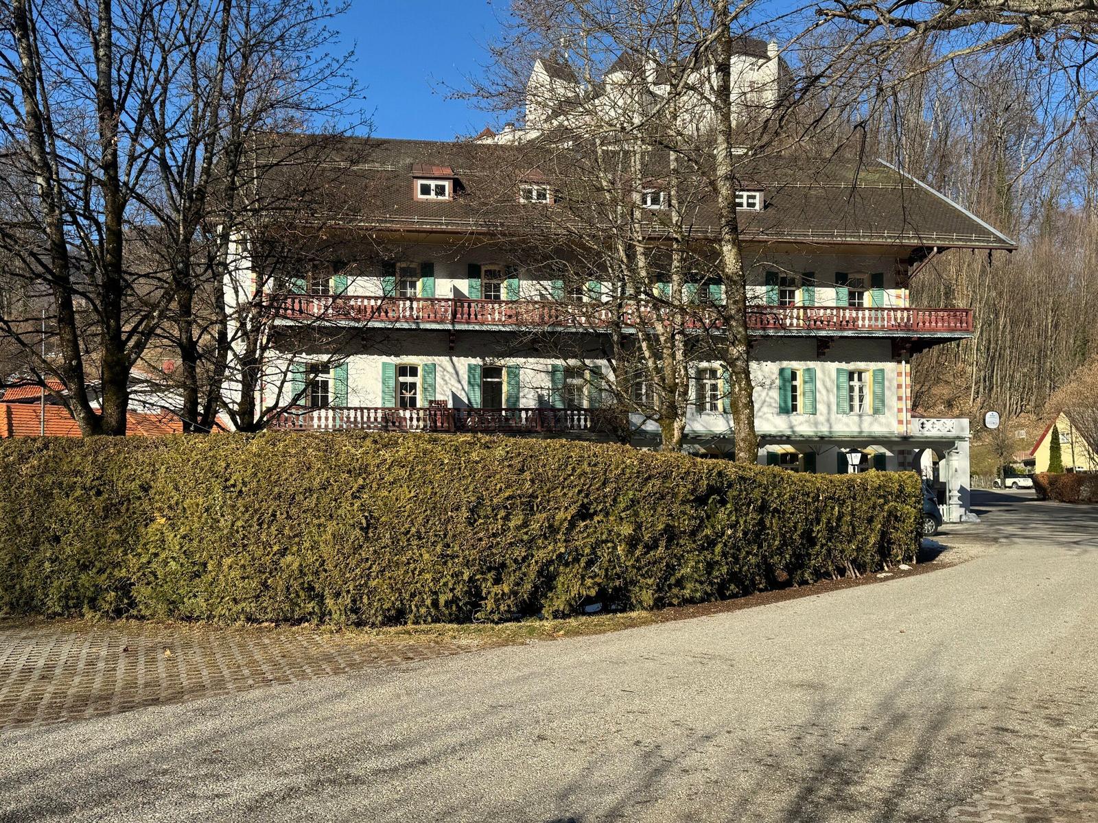 Ein großes Haus mit Balkonen und grünen Fensterläden, umgeben von Bäumen und einem Heckenzaun.
