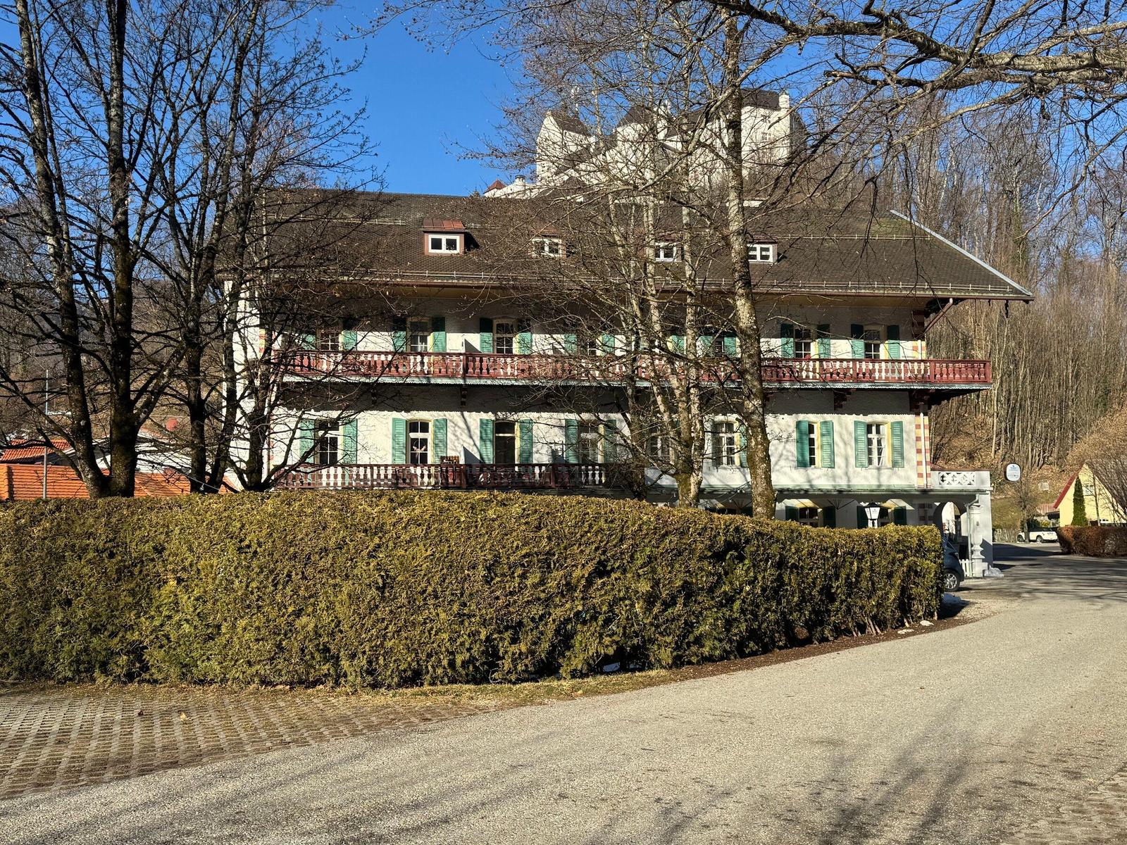 Haus mit Balkon, grünen Fensterläden und hohen Bäumen im Hintergrund.
