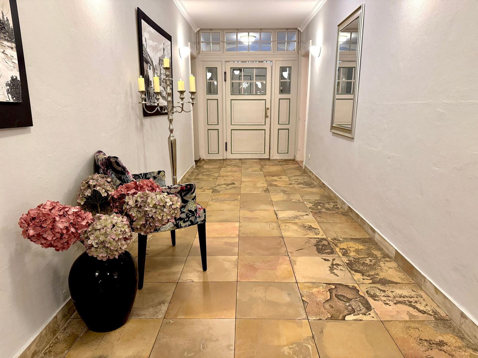 Hausflur mit Steinboden, Stuhl, Blumen und Wandspiegel. Tür mit Fenstern im Hintergrund.