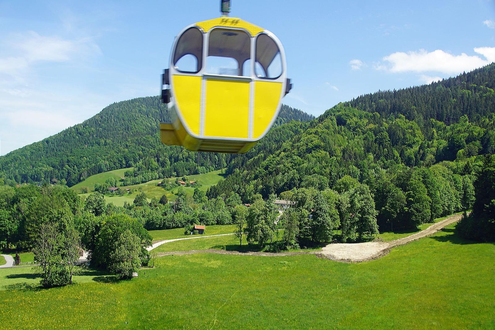 Gelber Seilbahnwagen schwebt über grüne Wiesen und bewaldete Berge.