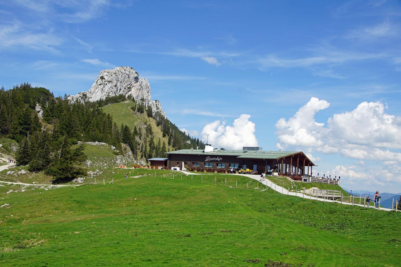 Hütte mit Terrasse auf grünem Hang, Berg im Hintergrund, blauer Himmel mit Wolken.