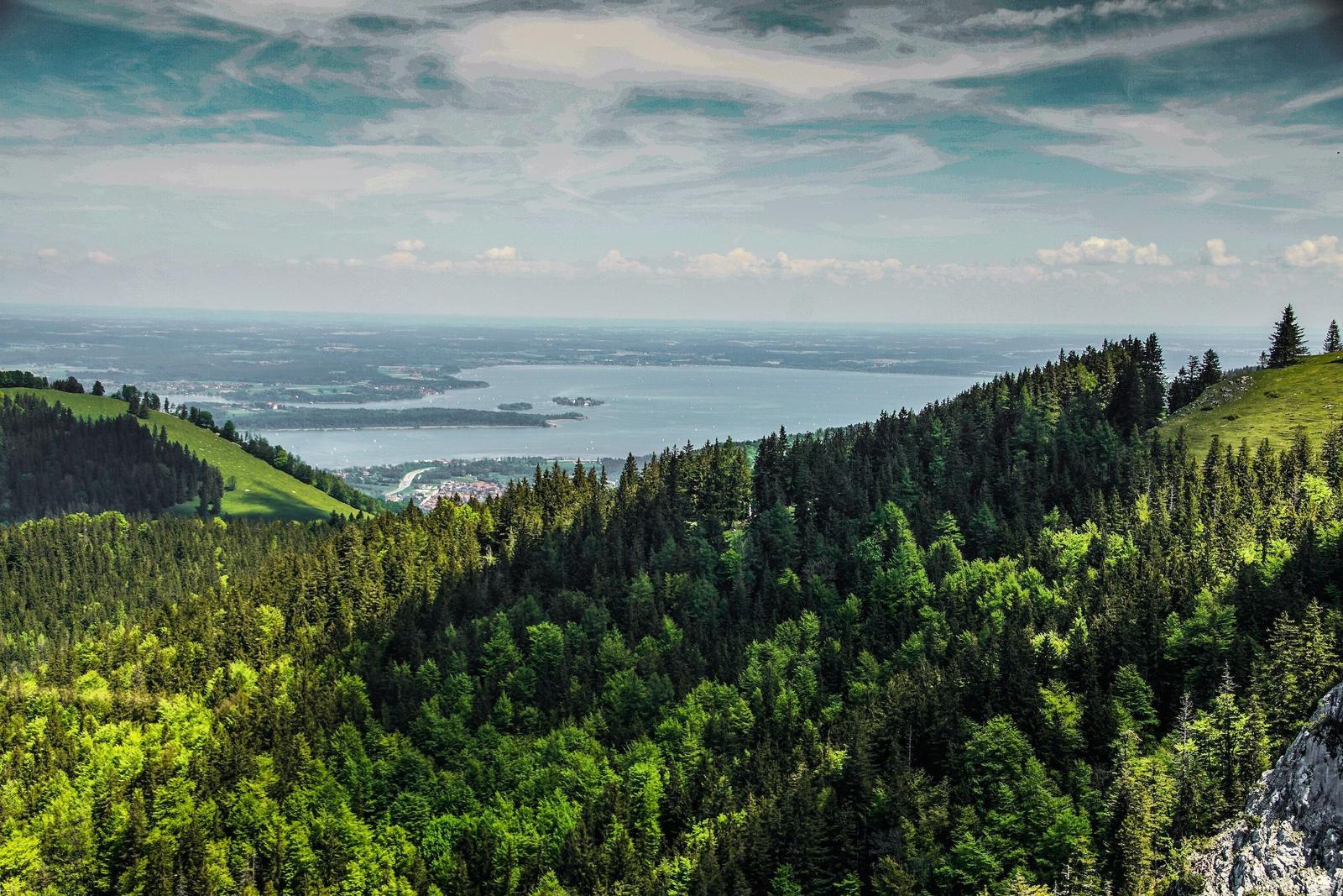 Panoramablick auf einen See, umgeben von Wäldern und Hügeln unter einem bewölkten Himmel.