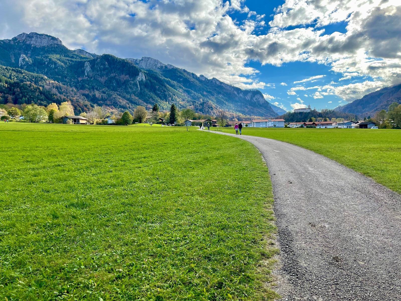 Gehweg durch grünes Feld mit Bergen und Dorf im Hintergrund.