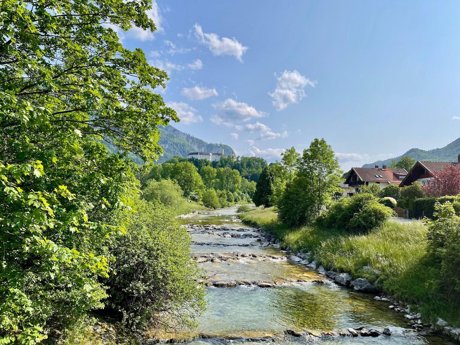 Fluss mit Steinen fließt durch grüne Landschaft mit Häusern und Schloss im Hintergrund.