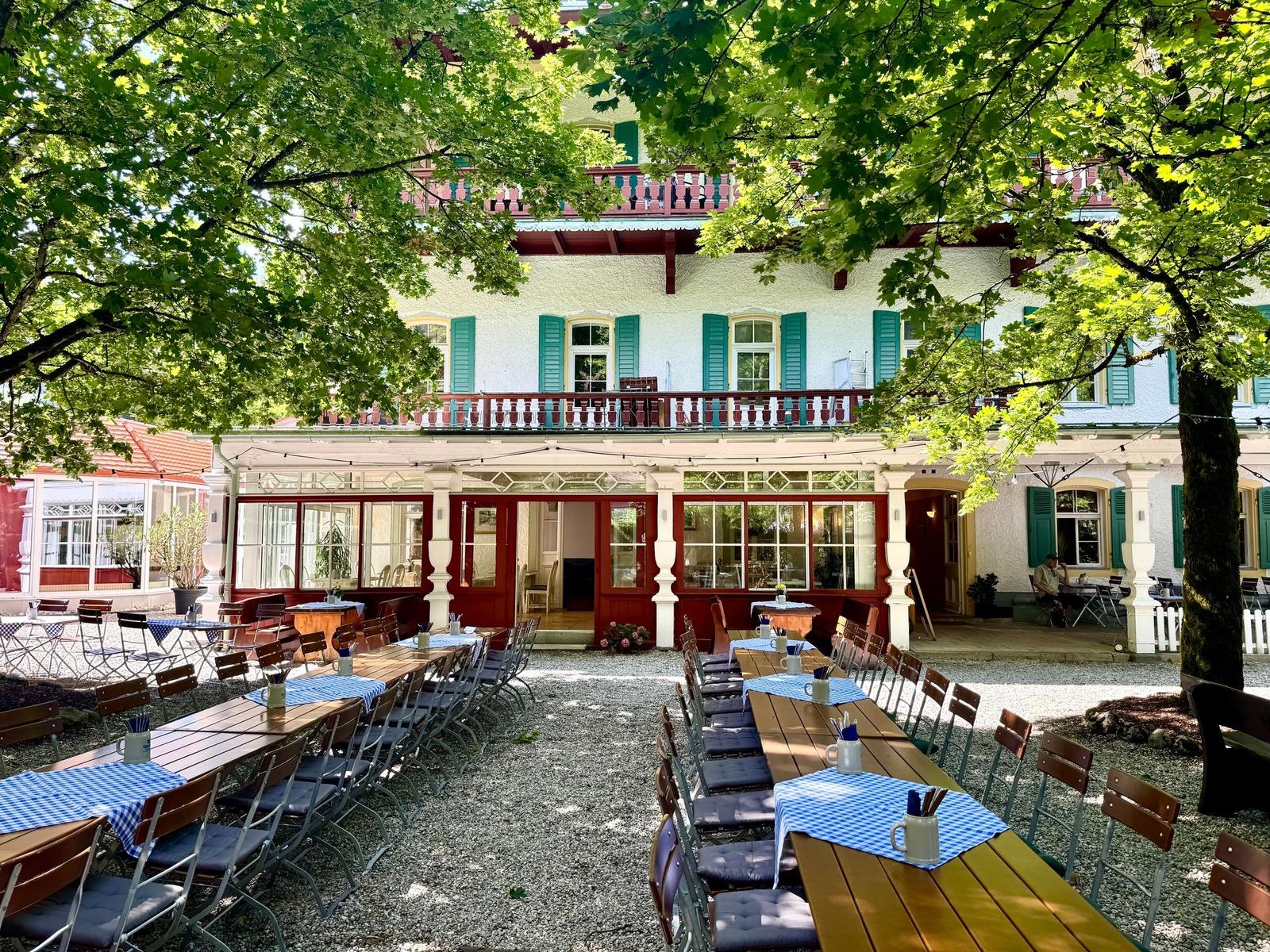 Hof mit Tischen und Stühlen unter Bäumen vor einem zweistöckigen Haus mit Balkon.