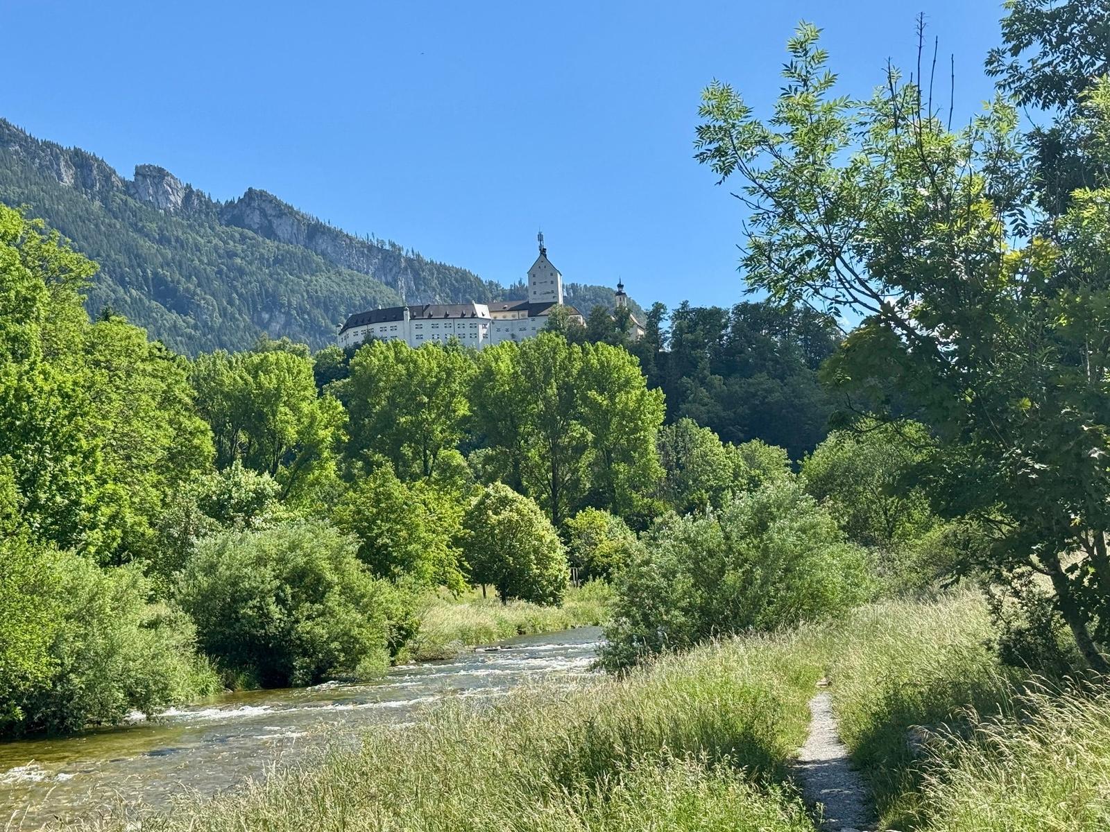 Flussuferweg mit Blick auf Schlossberg und bewaldete Hügel unter blauem Himmel.