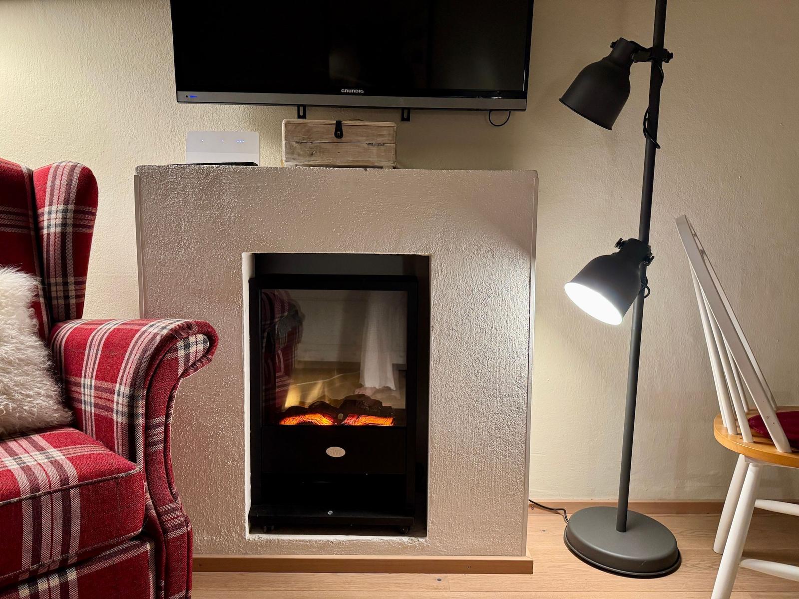 Ein Kamin mit brennendem Feuer, daneben ein roter Sessel und eine Stehlampe.