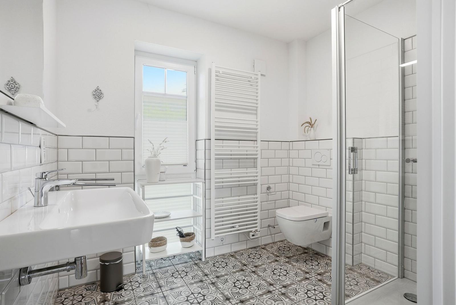 Badezimmer mit Dusche, Waschbecken und Toilette. Weiße Fliesen und gemusterte Bodenfliesen.