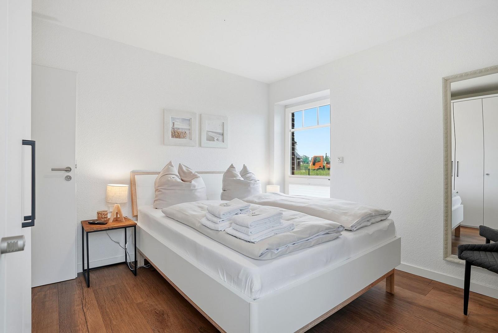 Schlafzimmer mit Bett, Nachttisch und Fenster. Spiegel und Holzboden sichtbar.